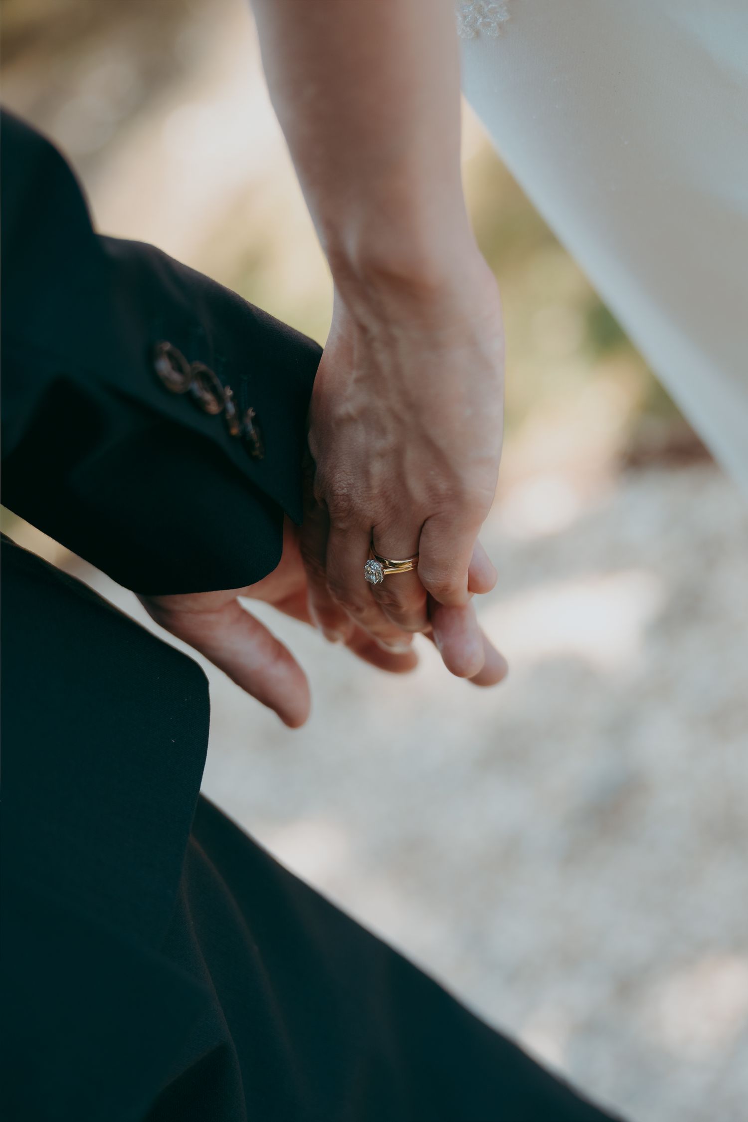 Close-up of a couple holding hands, bride wearing a gold diamond engagement ring, groom in dark suit.