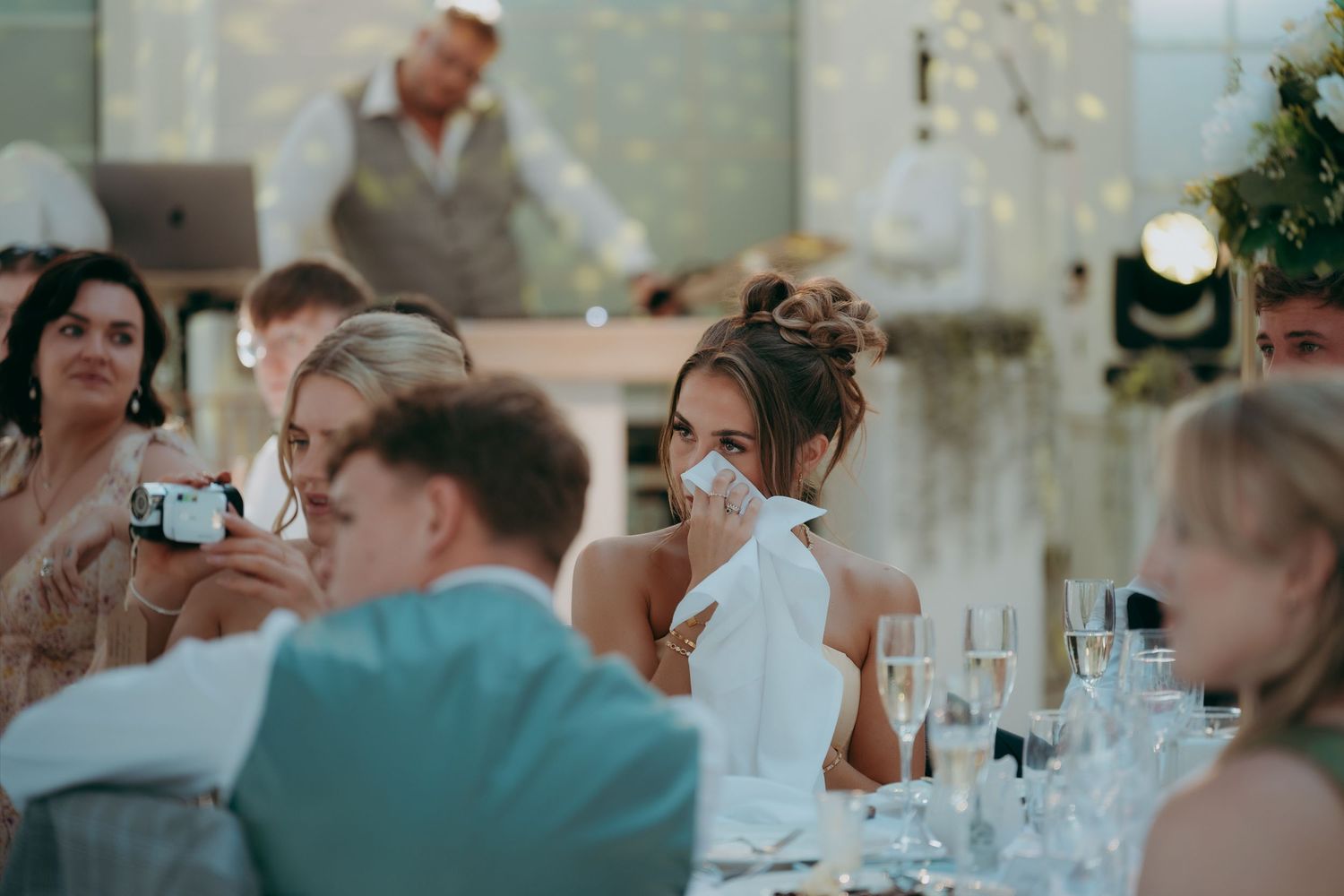 Emotional bride wiping tears during wedding reception speech at elegant venue with champagne glasses.