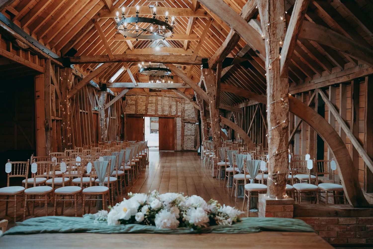 Rustic barn wedding venue with wooden beams, fairy lights, white floral centerpiece, and elegant chair-lined aisle.
