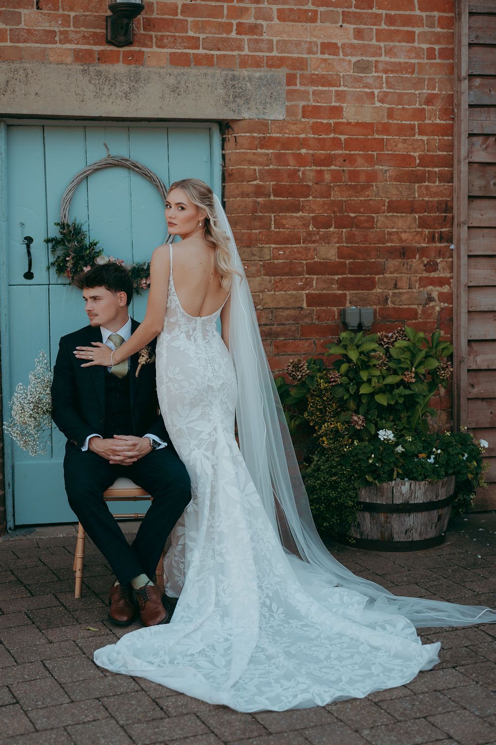 Bride in a lace backless wedding gown with a long veil stands beside a seated groom in a dark suit by a pale blue door and brick wall.