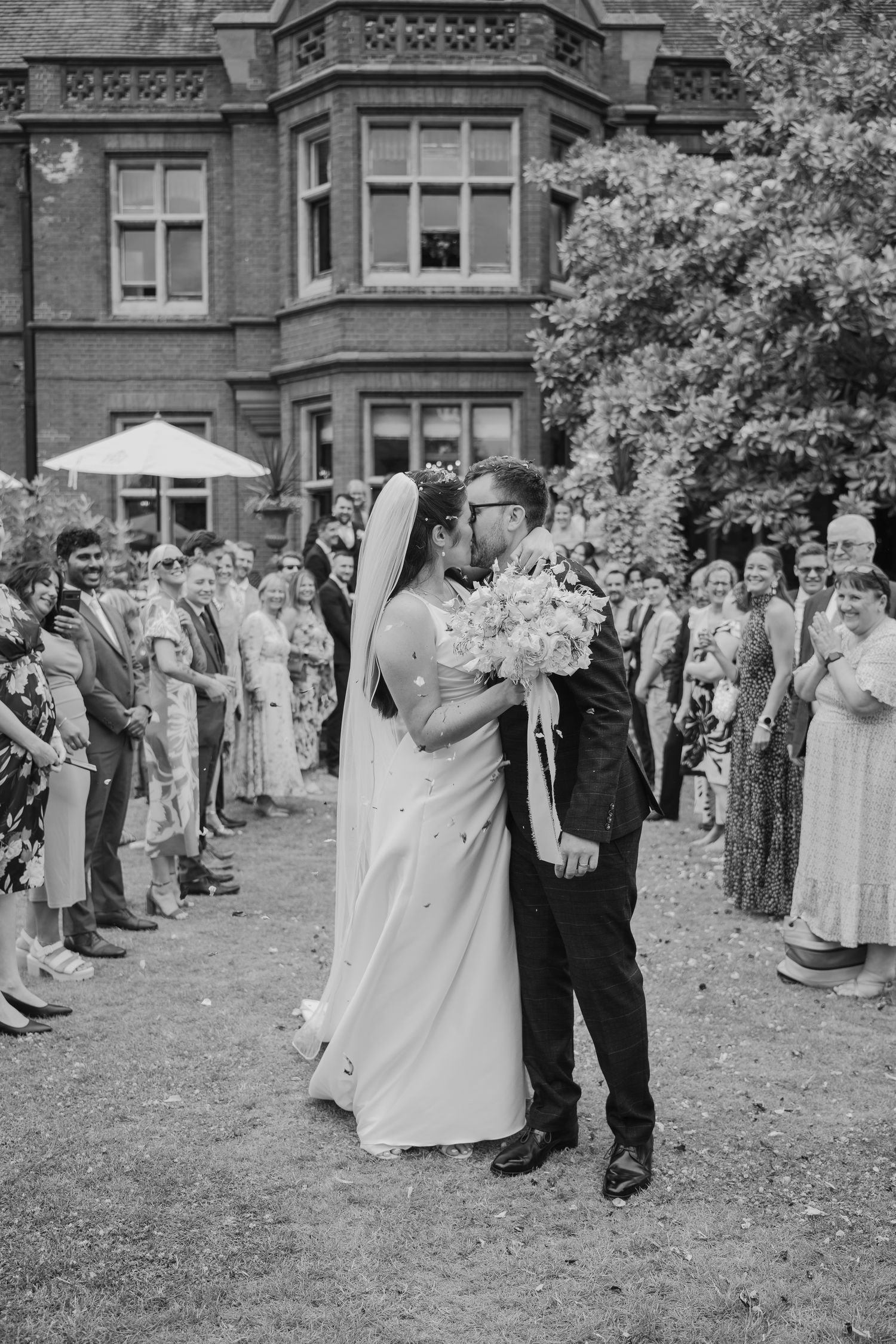 Black and white photo of a bride and groom sharing a kiss while guests line the path at an outdoor wedding venue.