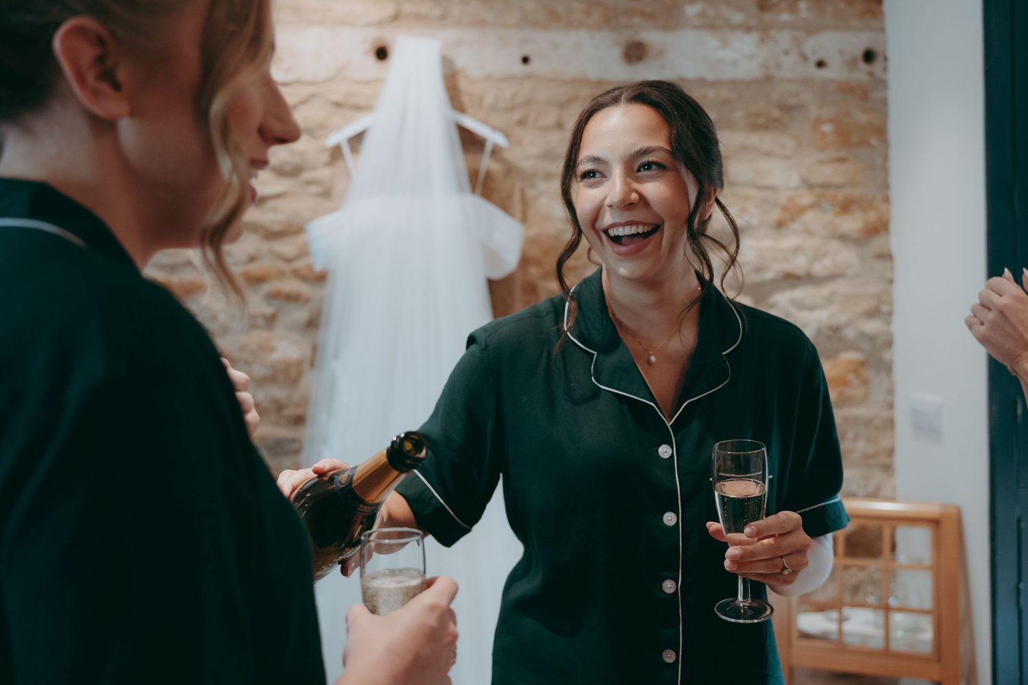 Two women in green pajamas celebrate with champagne near a wedding dress in a rustic stone-walled bridal suite.