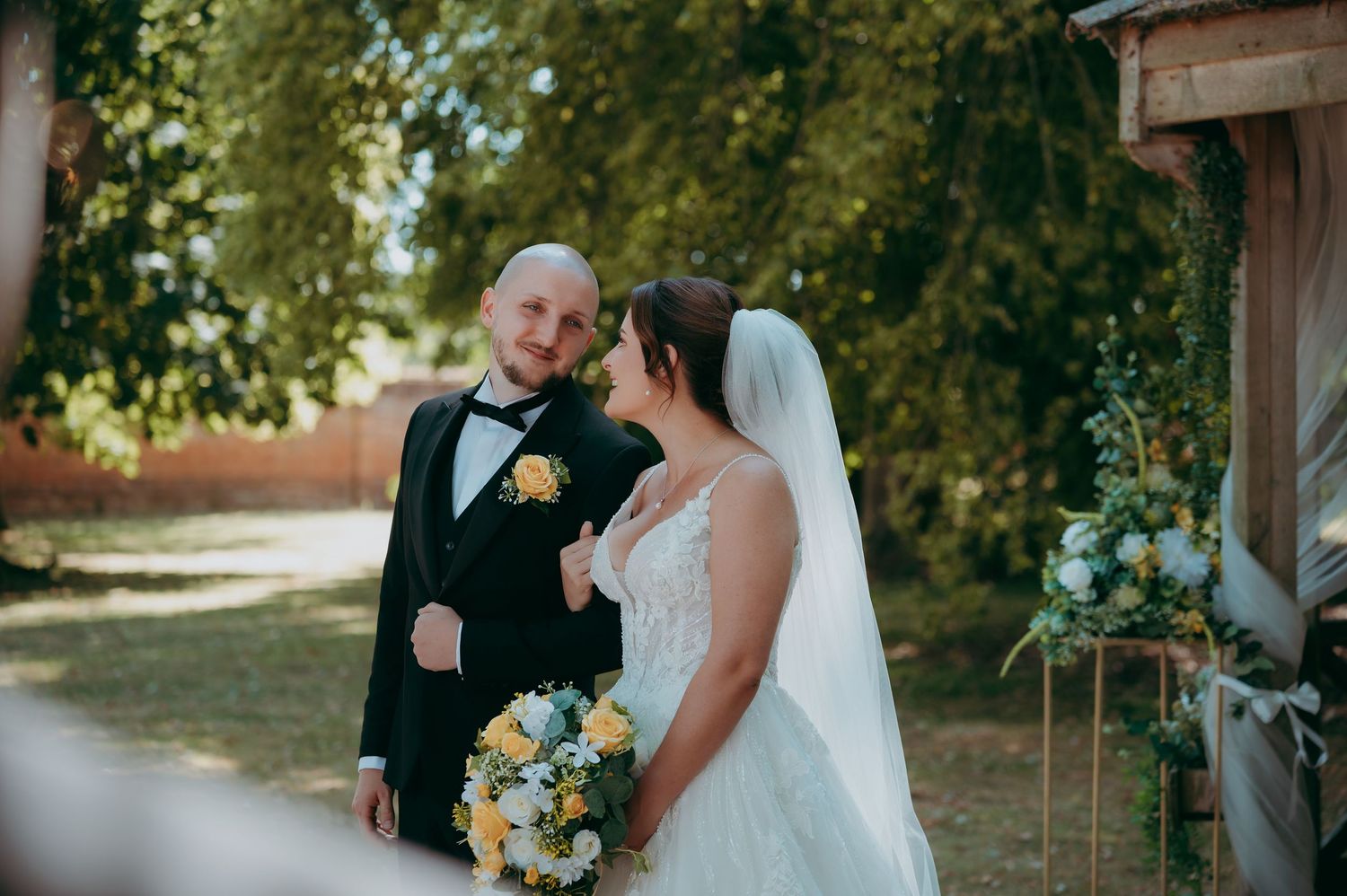 Bride and groom smiling at each other outdoors, bride holding a yellow and white bouquet in a lace wedding gown.