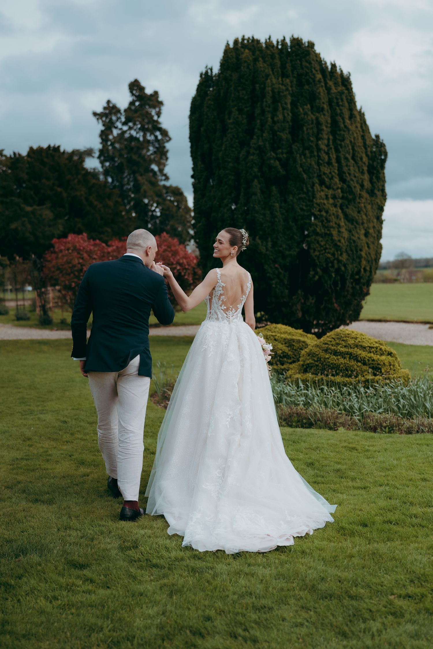 Bride in white lace gown and groom in dark suit walk hand in hand through lush garden on their wedding day.