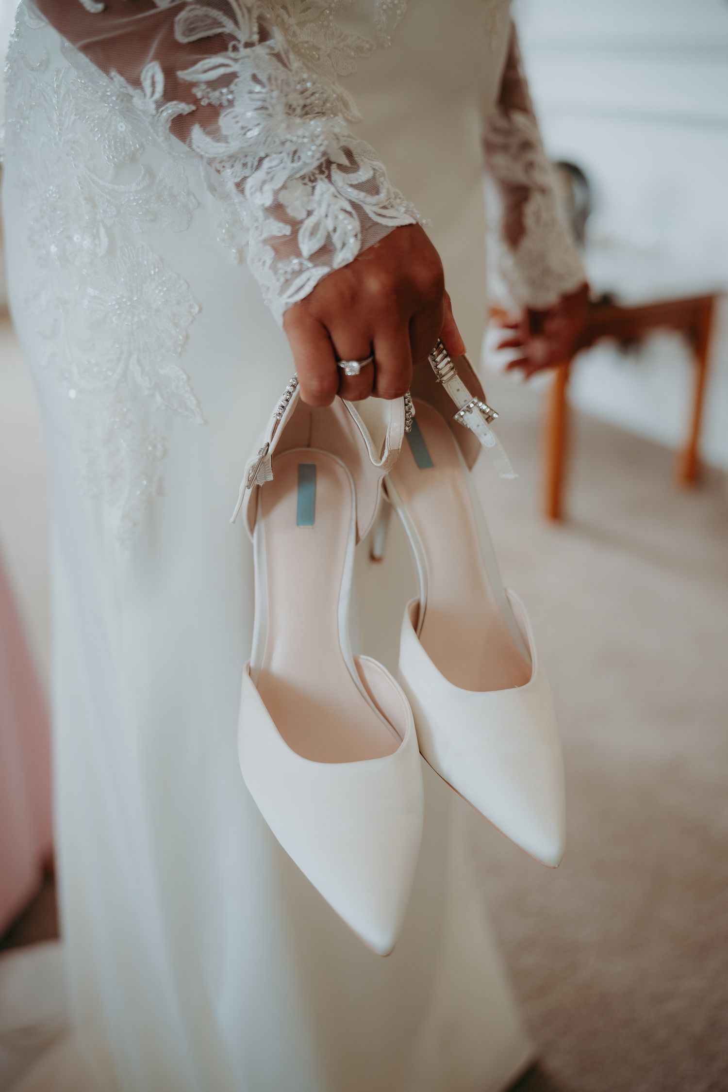 Bride in lace wedding gown holding elegant white pointed-toe heels with blue sole detail and diamond rings on her hand.