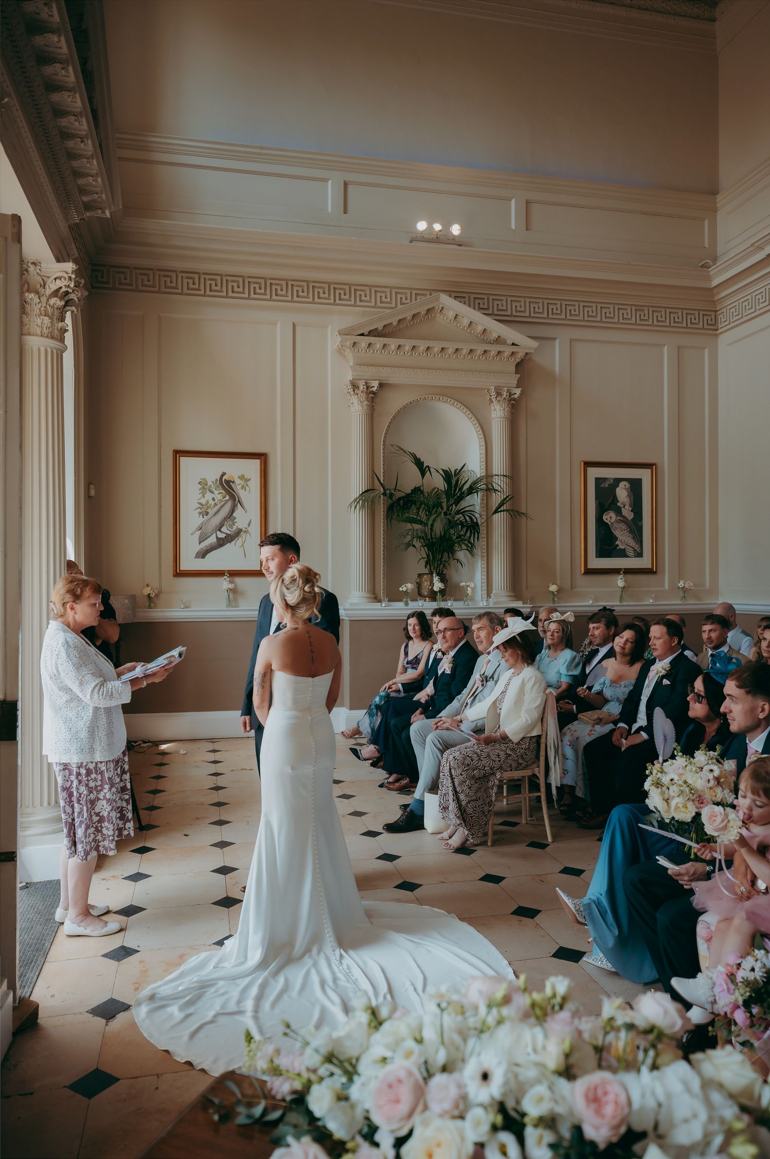 Elegant wedding ceremony in a grand hall with bride and groom at the altar, surrounded by seated guests and floral arrangements.
