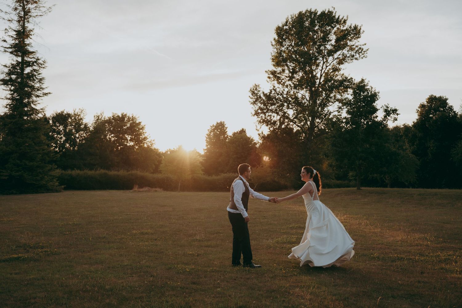 Bride and groom dance hand in hand in a sunlit field at golden hour with trees in the background.