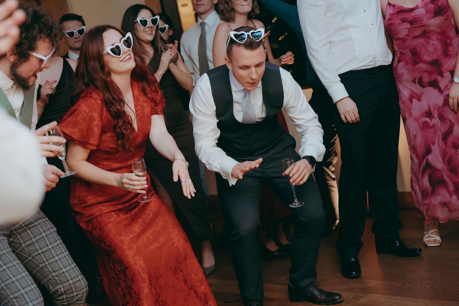 Guests dancing energetically at a wedding reception, woman in red dress and man in white shirt with novelty sunglasses.