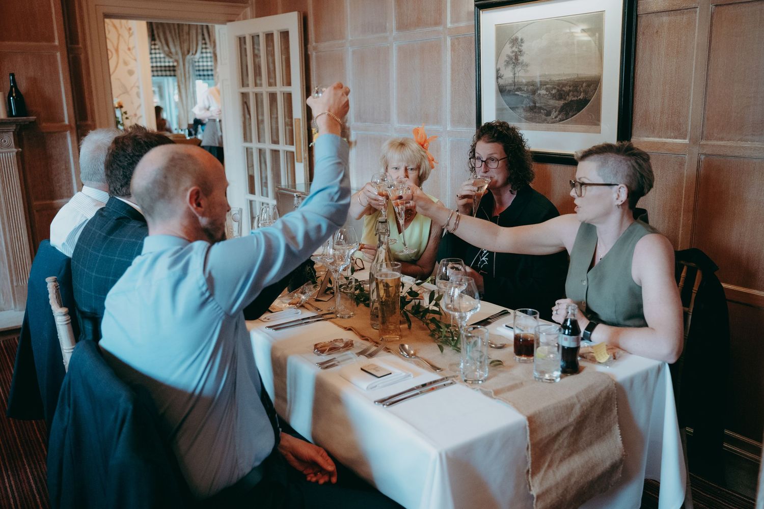 Guests raising glasses in a toast at a wedding reception dinner table in an elegant wood-paneled restaurant.