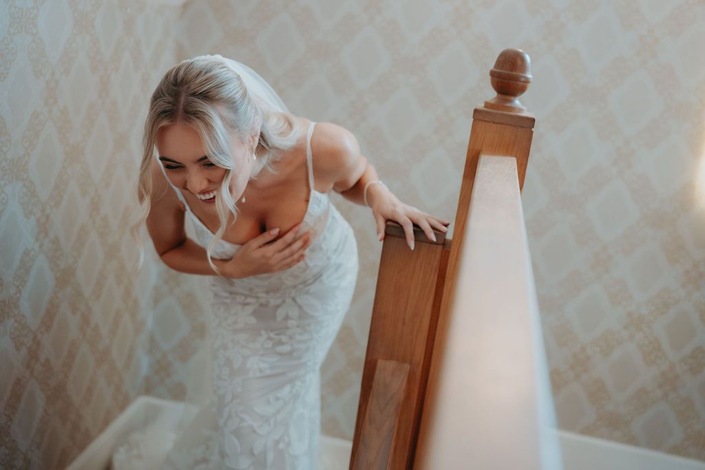 Smiling bride in a white lace wedding dress leans over a wooden staircase banner in a softly lit room with patterned wallpaper.