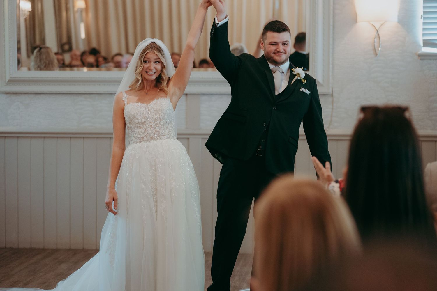 Bride and groom celebrate their first dance at wedding reception, raising hands joyfully in elegant white venue.
