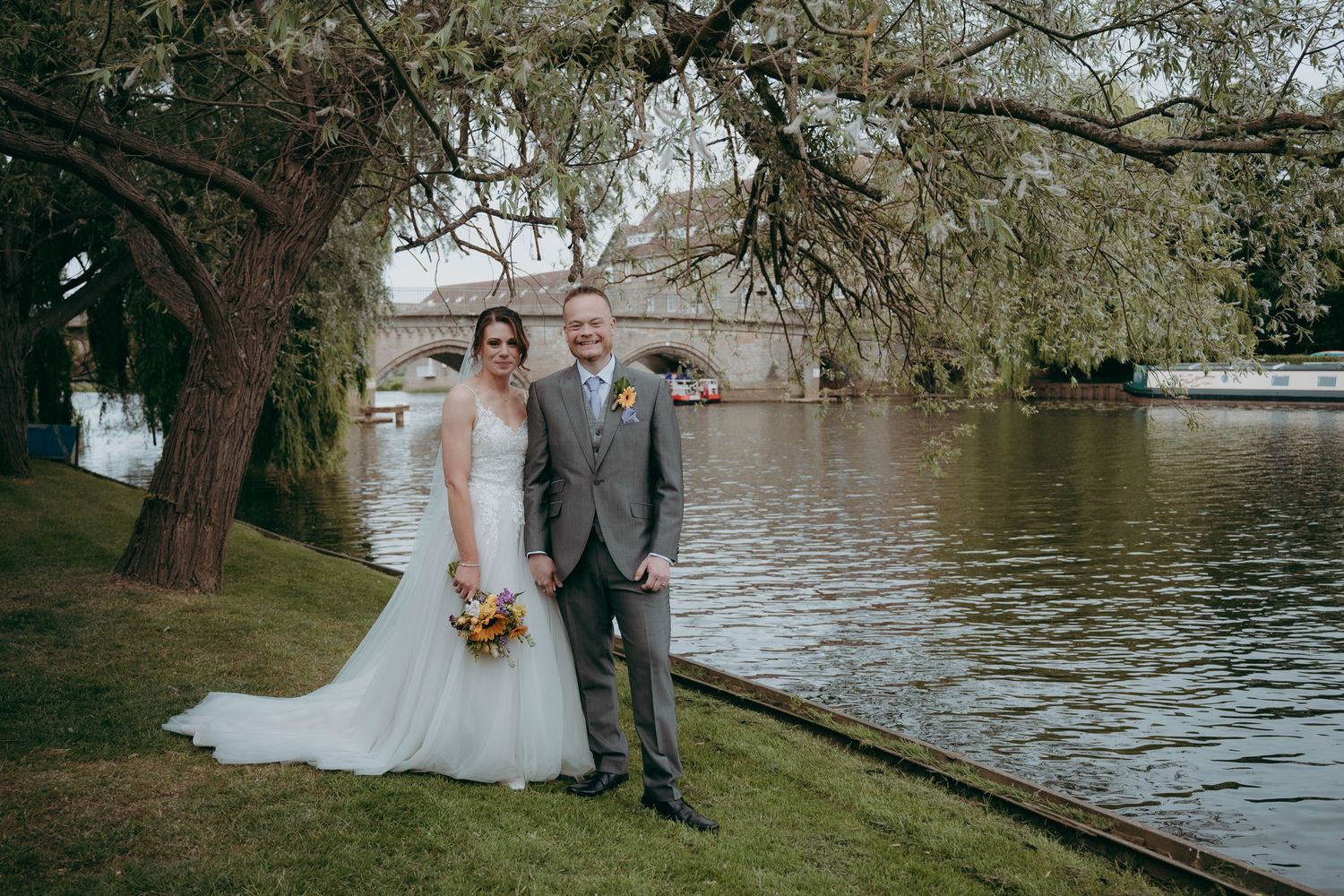 Bride and groom posing by a river under willow trees, bride in white gown holding bouquet, groom in grey suit.