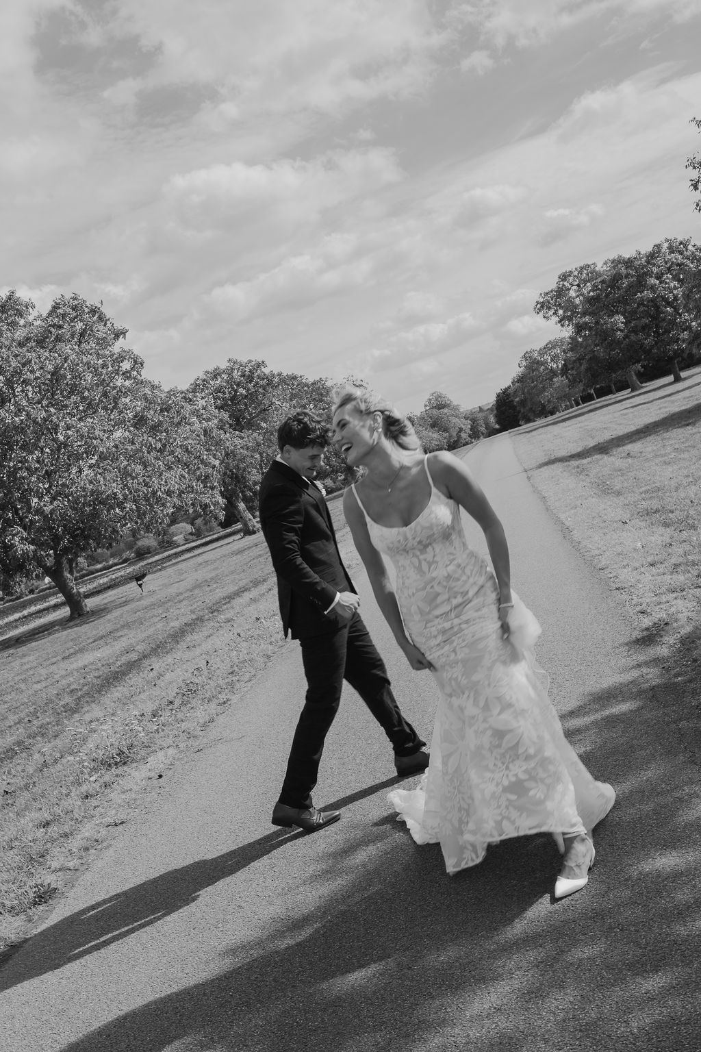 Black and white photo of a bride in a lace gown and a groom in a suit laughing as they walk down a path in a sunlit park.