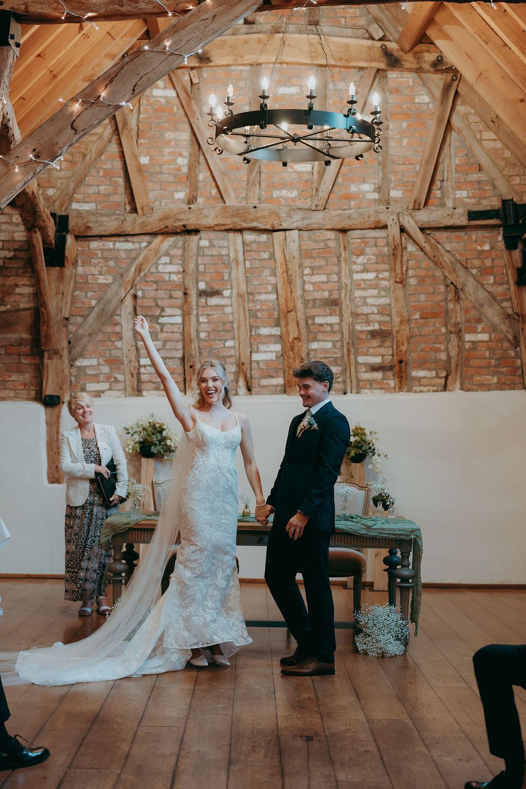 Bride in a lace gown raises her arm in joy beside her groom during a rustic wedding ceremony in a timber-framed barn venue.