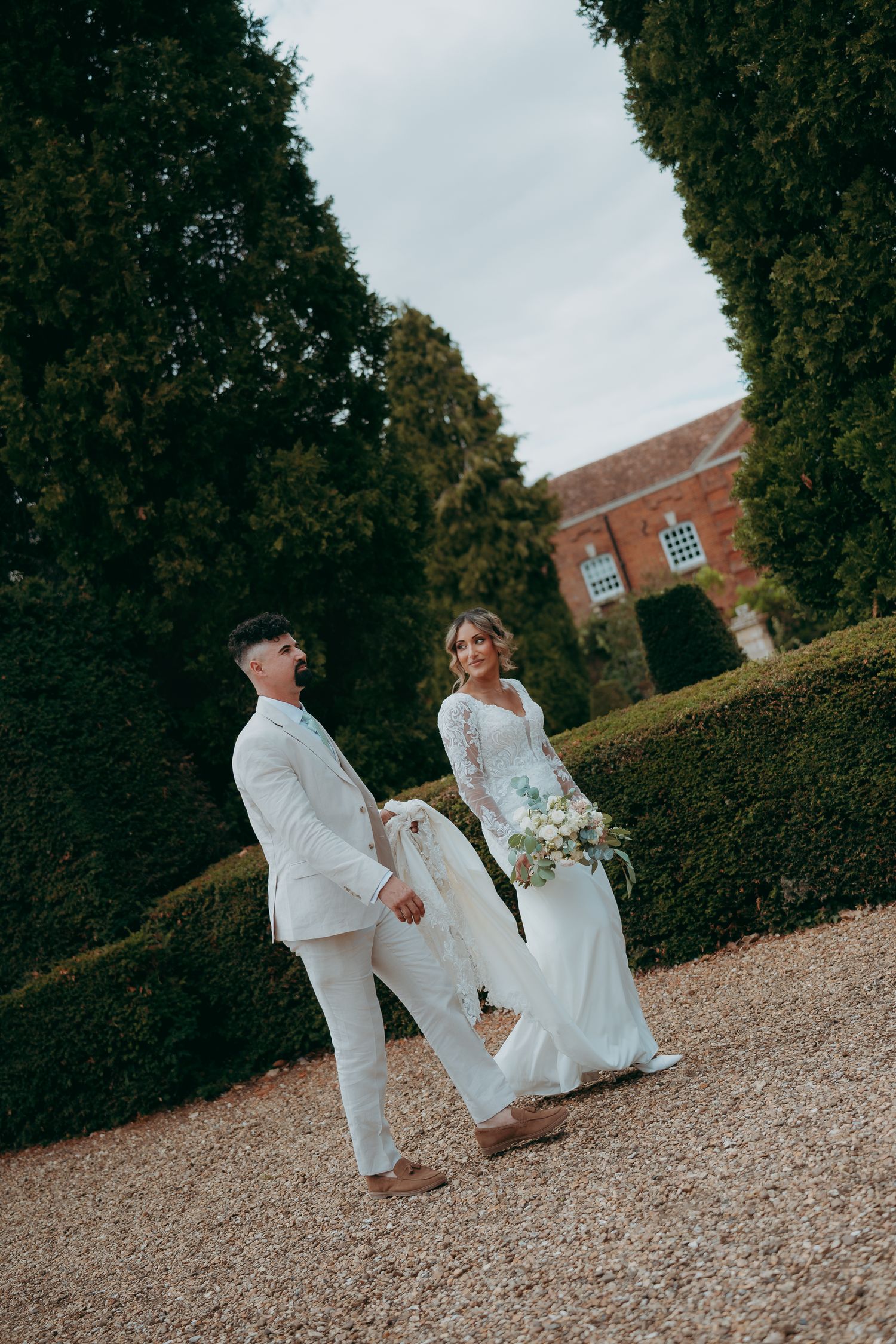 Bride and groom walking hand in hand on gravel path in lush garden, bride holding bouquet in elegant white gown.