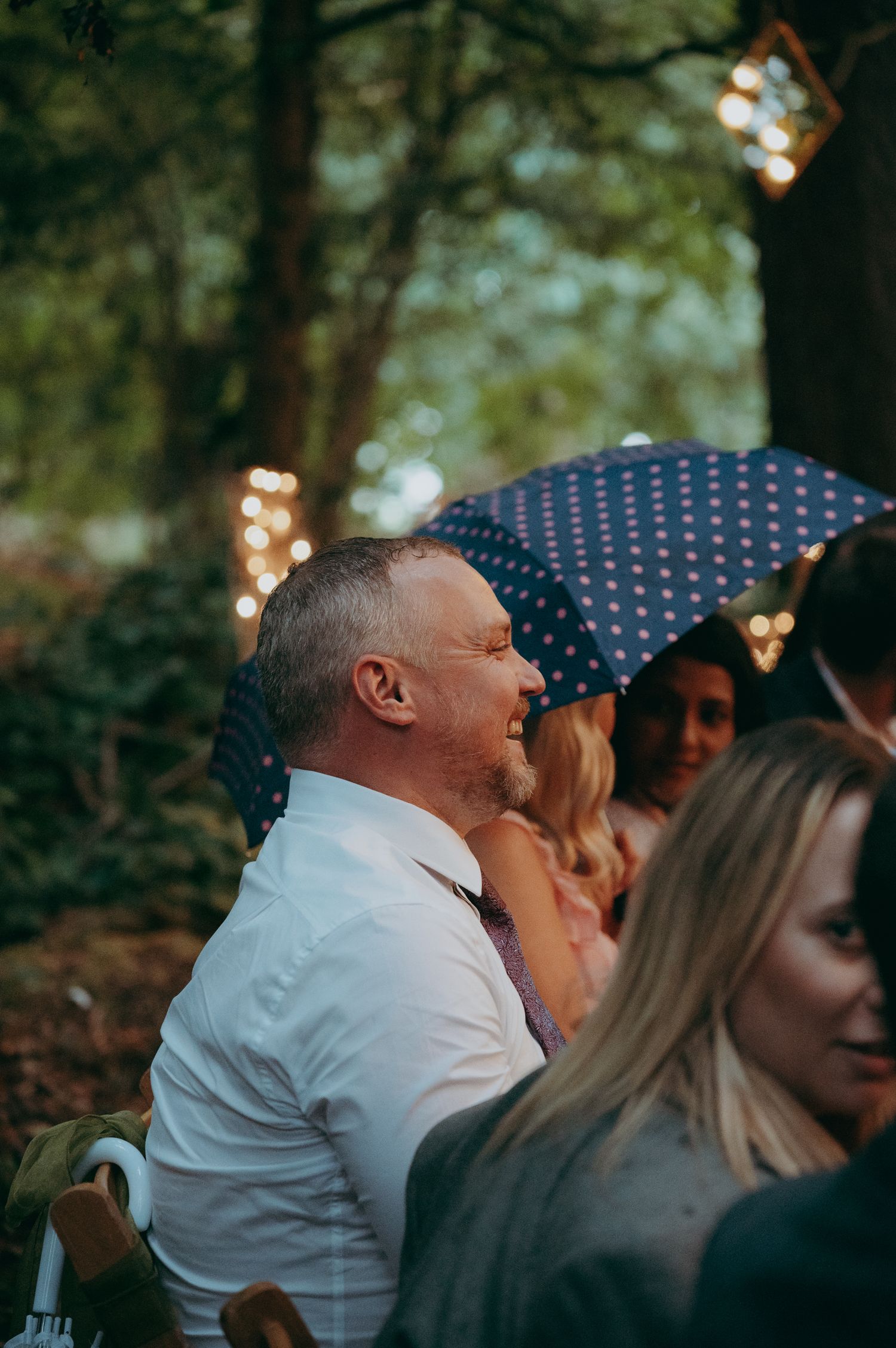 Man in white shirt smiling at outdoor woodland wedding, guests with polka dot umbrella and fairy lights in background.