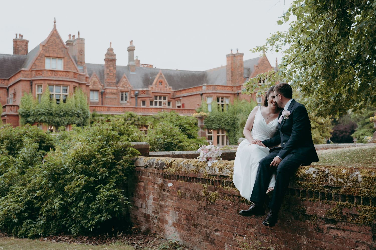 A bride and groom sit together on a brick wall in lush gardens outside a grand red-brick English manor on their wedding day.