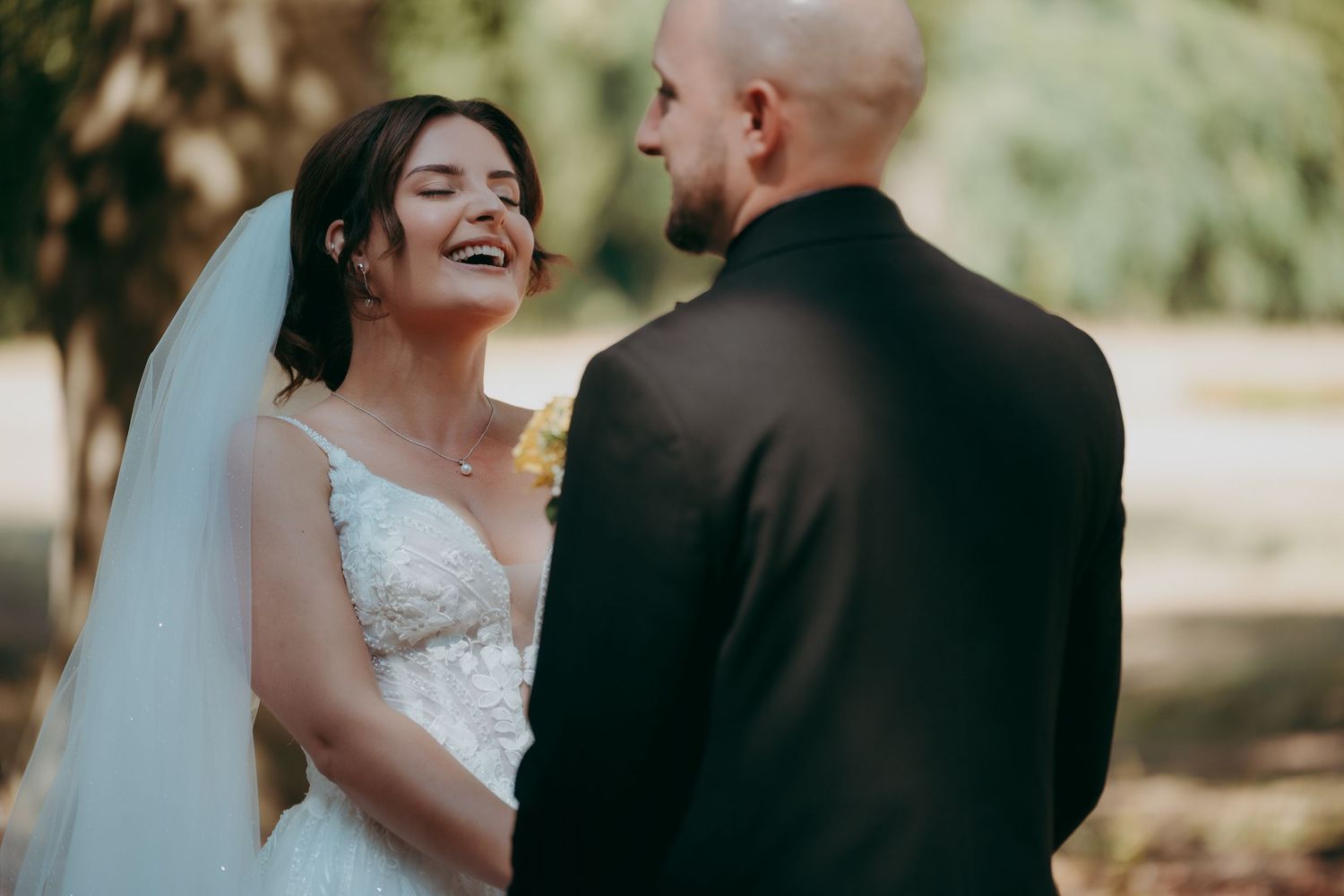 Bride in lace wedding dress laughing joyfully with groom in black suit during outdoor wedding ceremony.