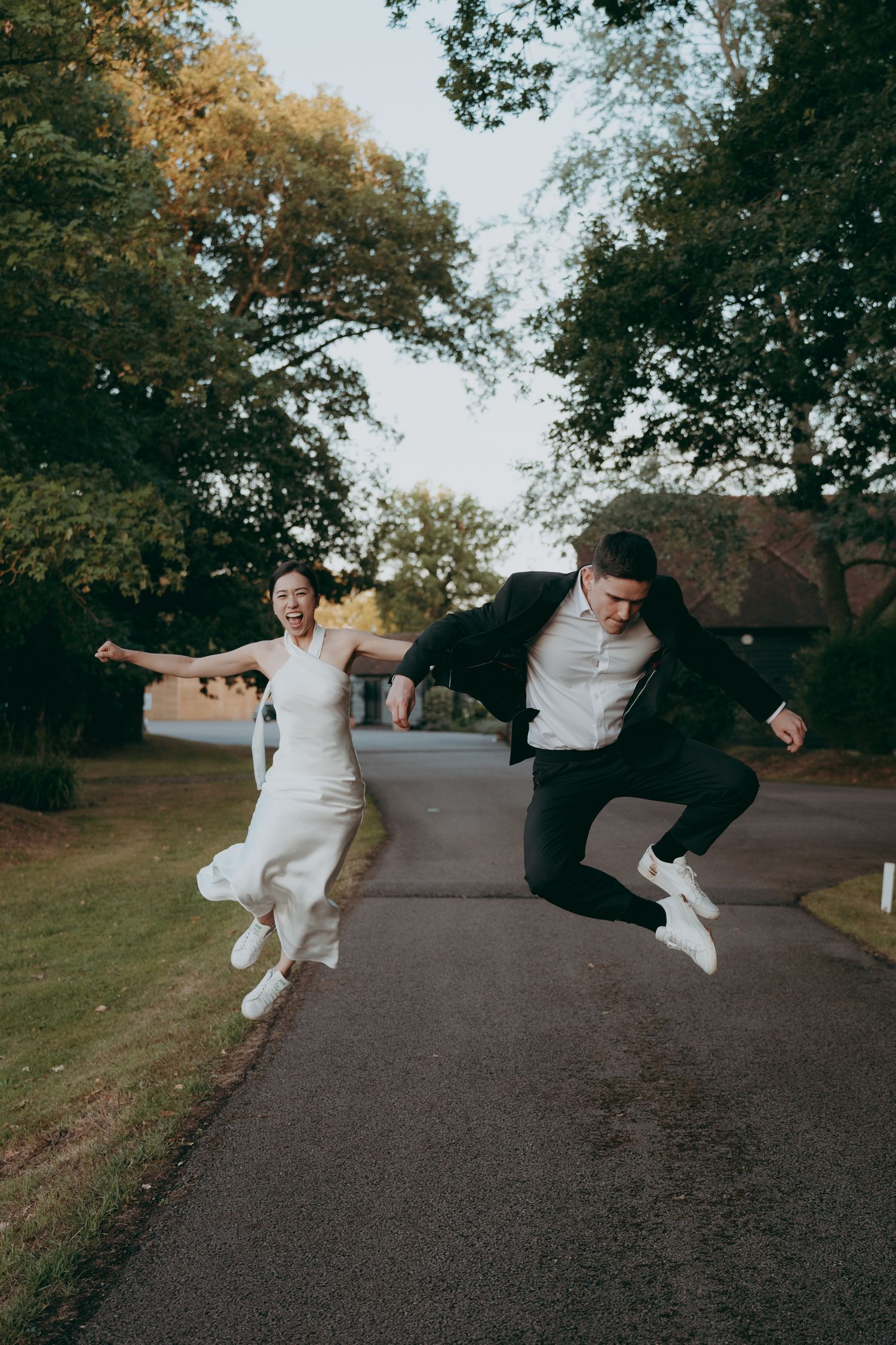 Joyful newlyweds in wedding attire jumping together on a tree-lined path, bride in white dress and groom in black suit.
