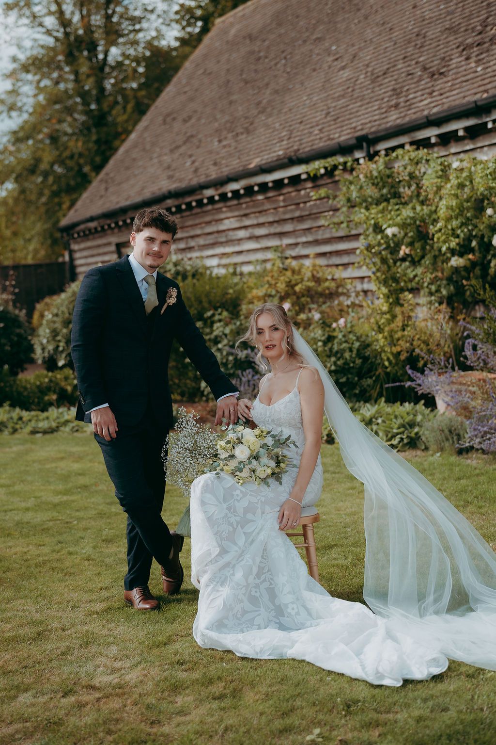 Bride in a lace wedding gown and long veil sits holding a bouquet beside her groom in a navy suit on a lawn before a rustic barn.