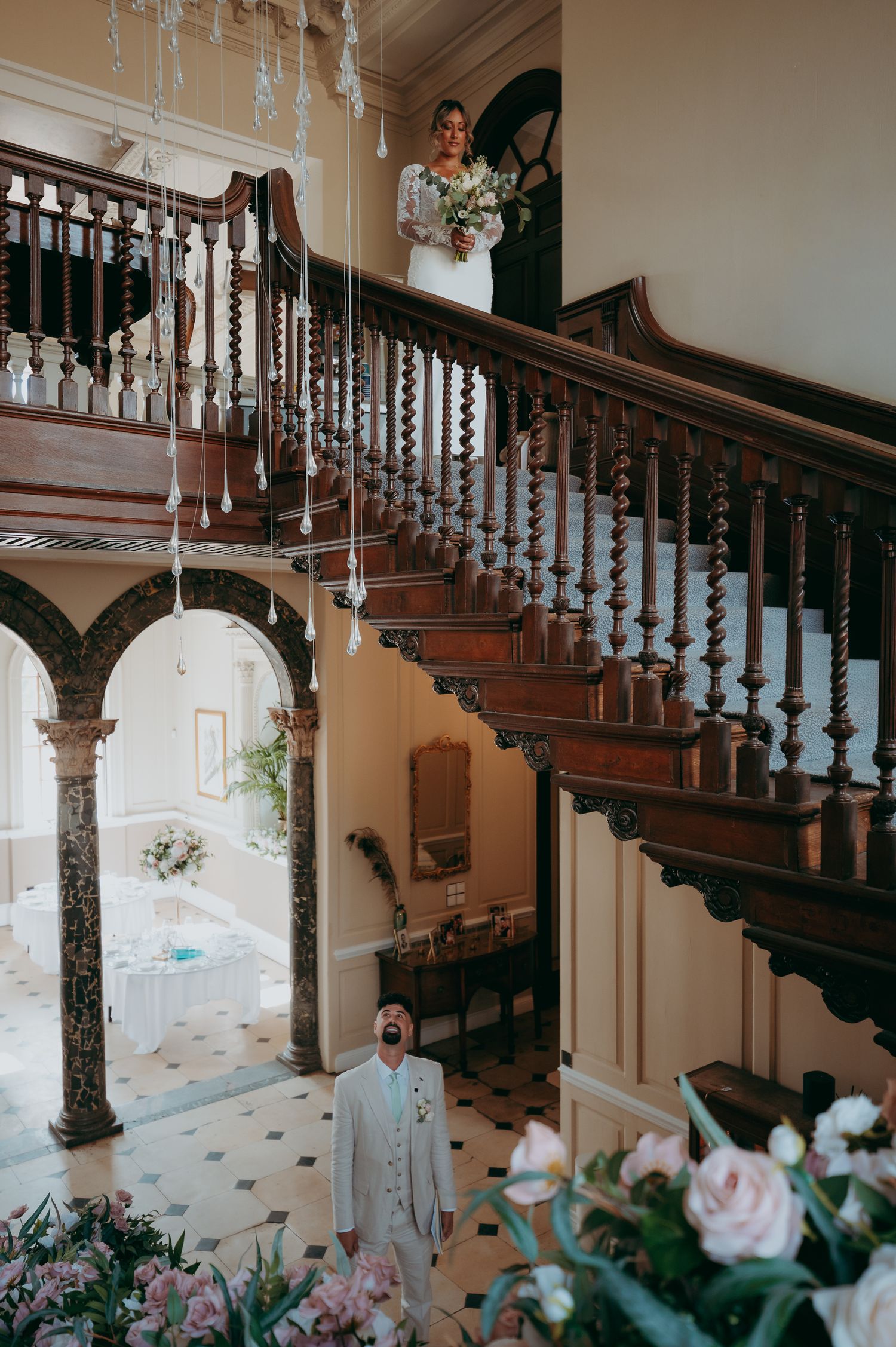 Groom looks up in awe at bride descending grand wooden staircase in elegant mansion wedding venue.