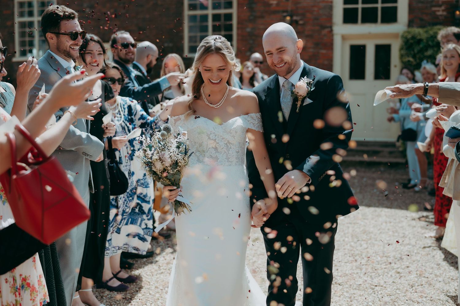 Bride and groom walk through confetti shower surrounded by cheering guests outside a brick venue on their wedding day.