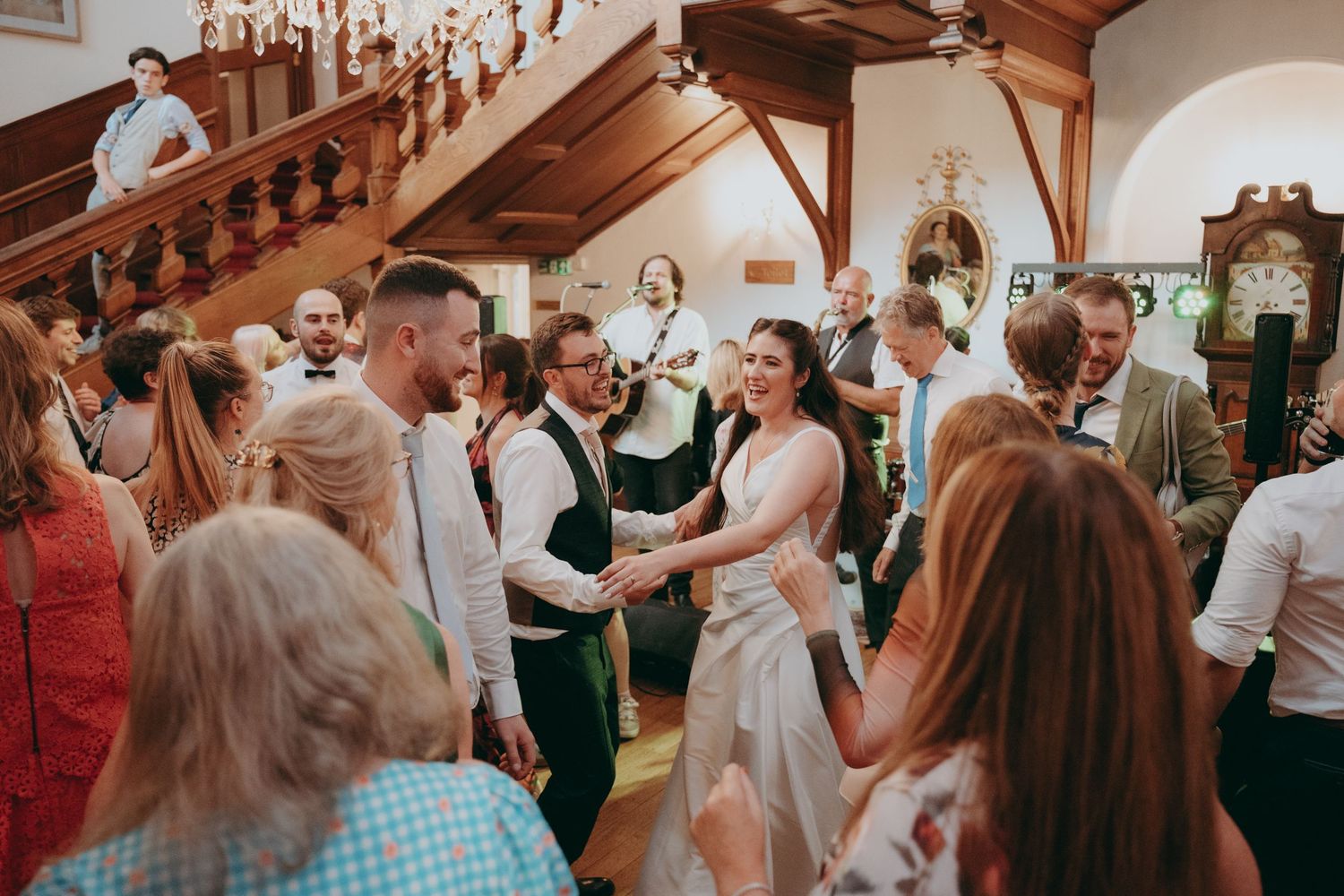 Bride and groom shaking hands with wedding guests in elegant venue with grand staircase during reception celebration.