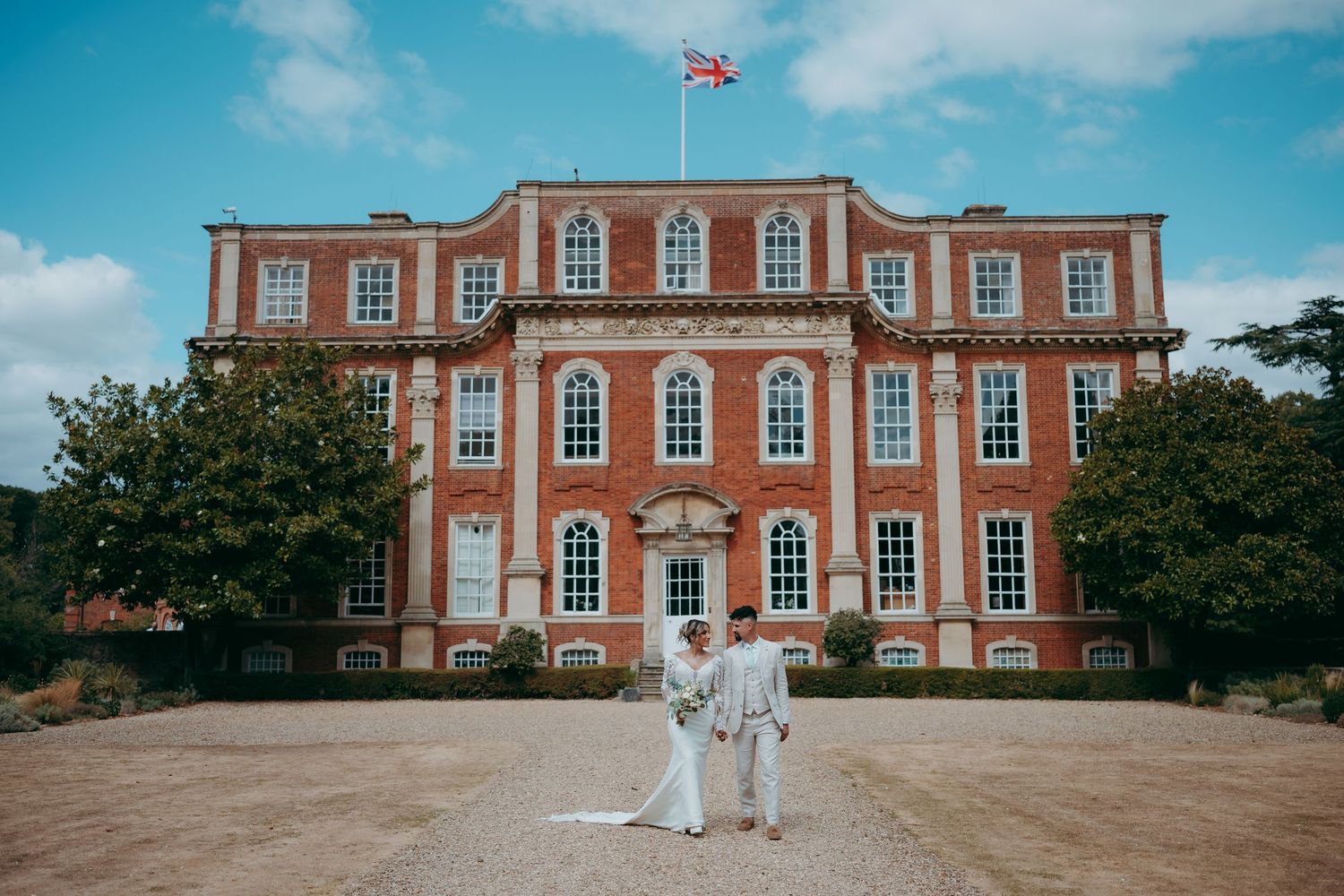 Bride and groom pose in front of a grand red brick Georgian manor with Union Jack flag, under blue skies.