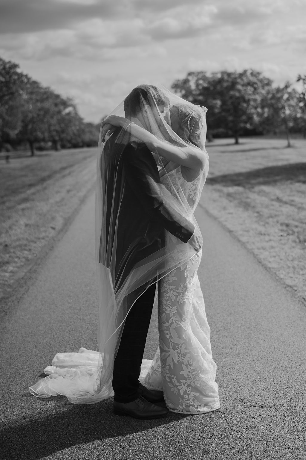 Bride and groom embrace under a sheer wedding veil on a tree-lined country road in a black and white wedding photograph.