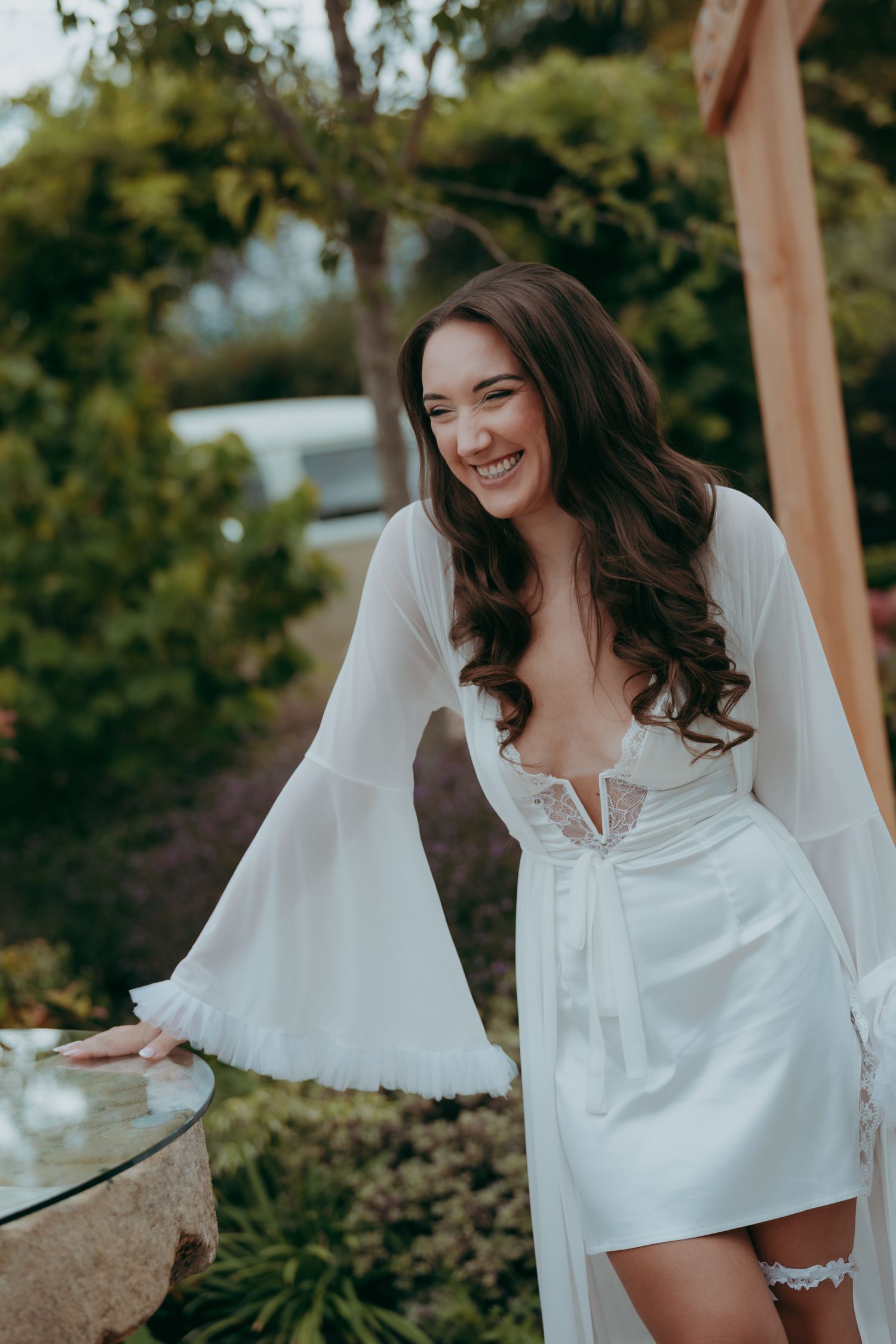 Smiling bride in white lace robe posing outdoors in a garden setting on her wedding day.