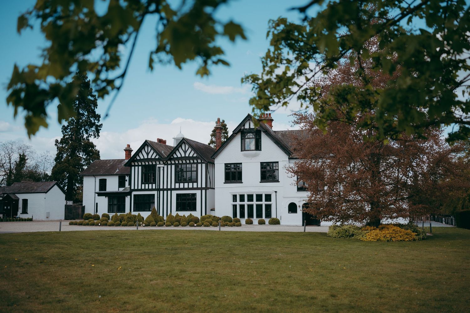 Tudor-style country manor house with black and white timber framing, surrounded by manicured lawns and autumn trees.