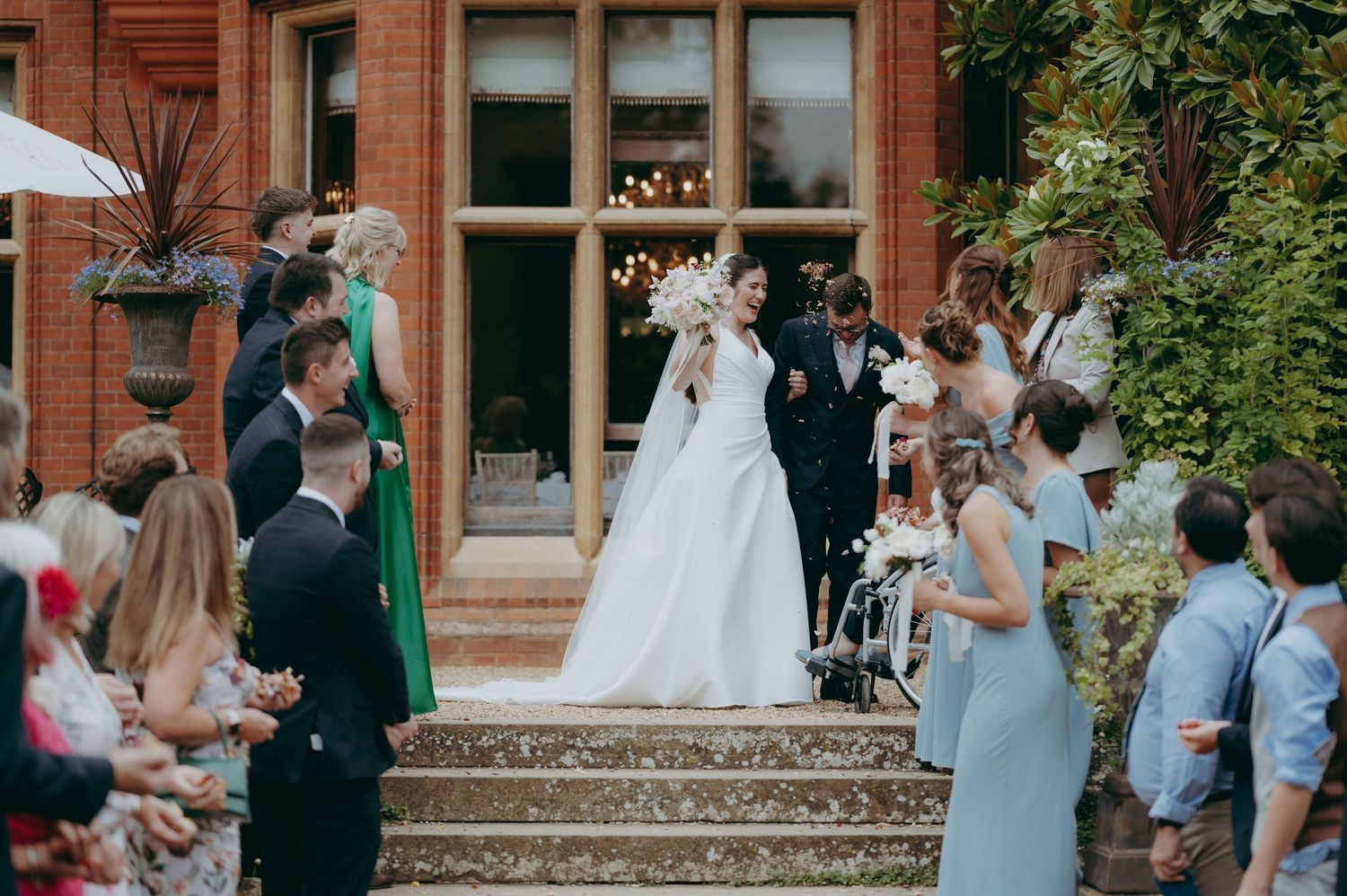 Bride and groom descend steps at elegant brick venue, greeted by guests throwing confetti at outdoor wedding ceremony.