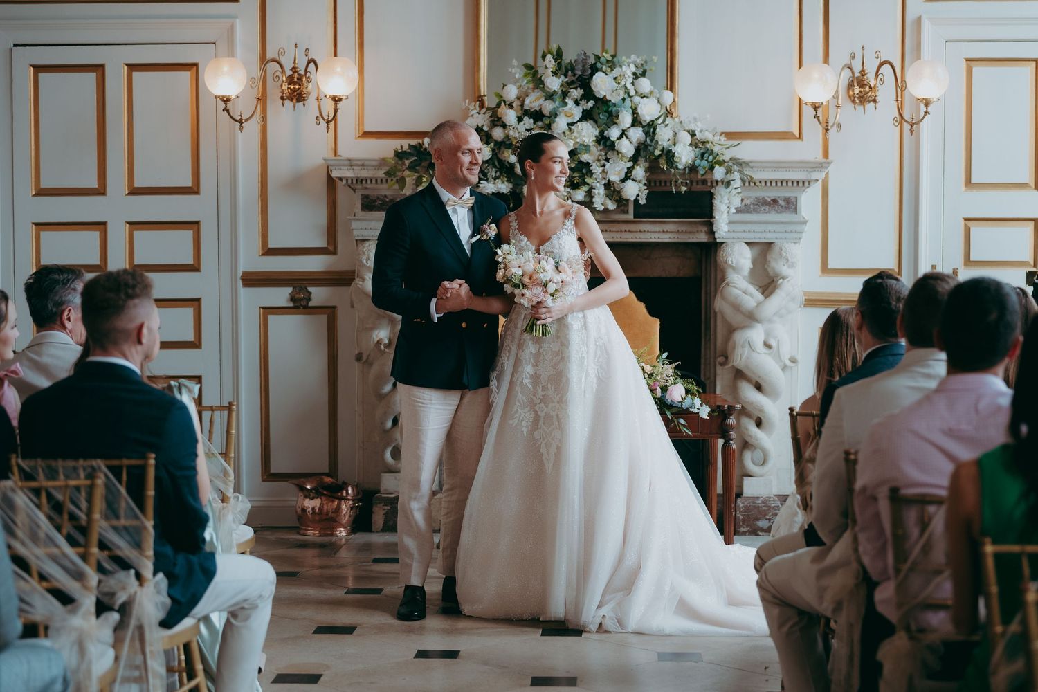 Bride in white gown walks down aisle with escort in elegant venue with floral arch and seated wedding guests.