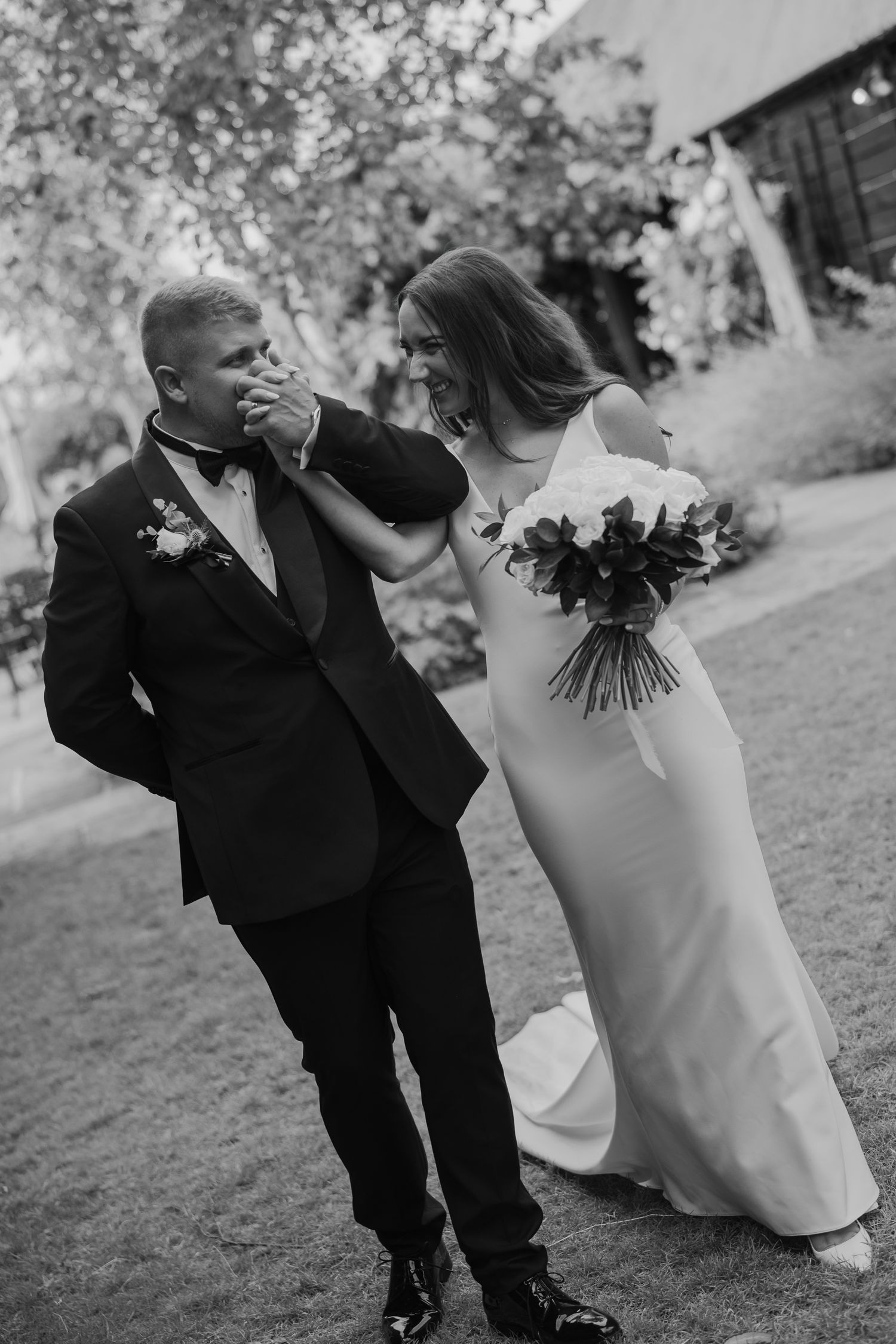 Black and white photo of a bride and groom laughing outdoors, she holds bouquet while he kisses her hand in formal attire.