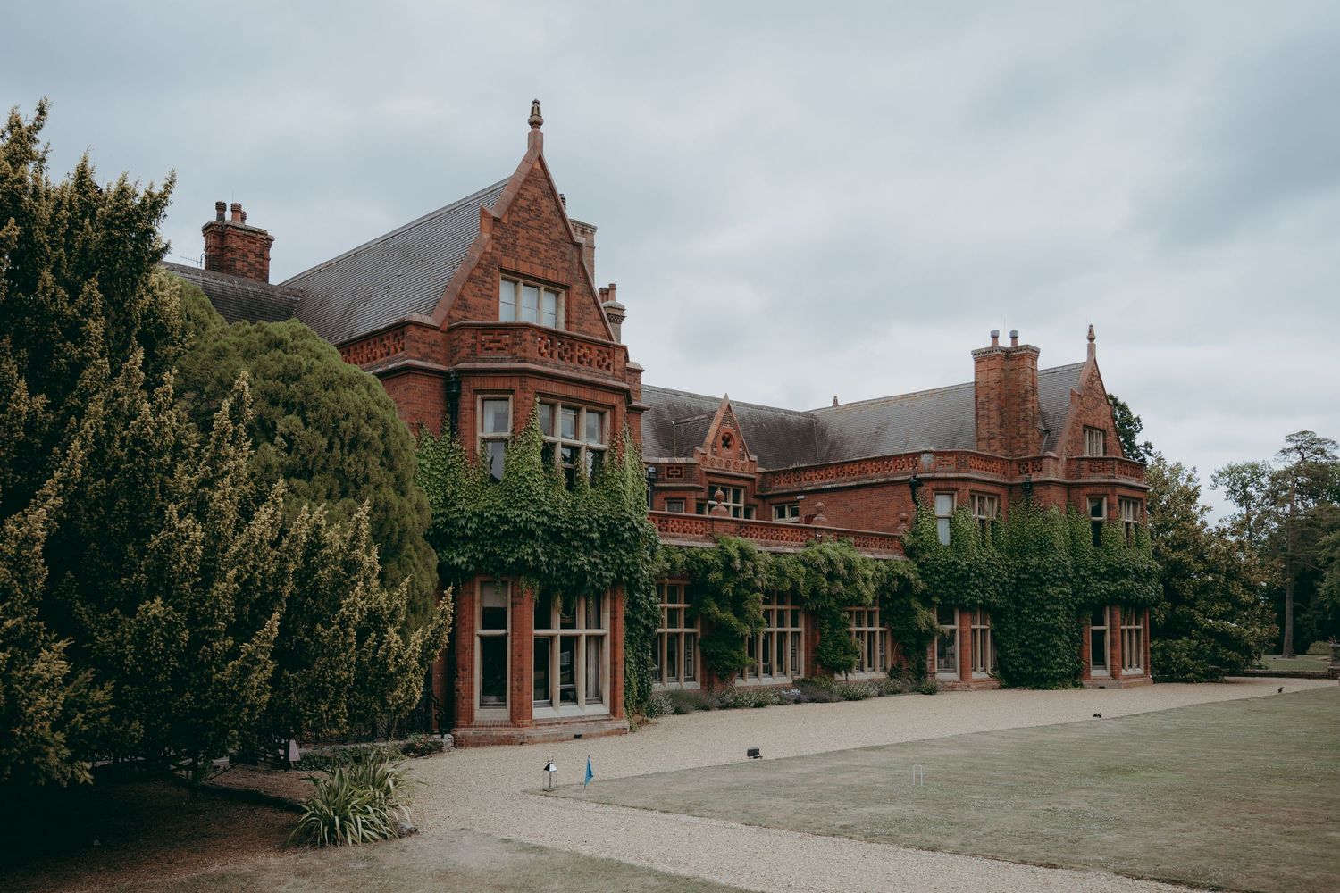 Grand red brick English manor house with ivy-covered walls, gabled rooflines, and manicured lawns under overcast skies.