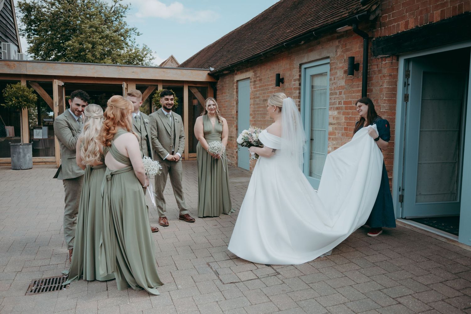 Bride in white gown with bridesmaids in sage green dresses outside a rustic brick venue on a wedding day.