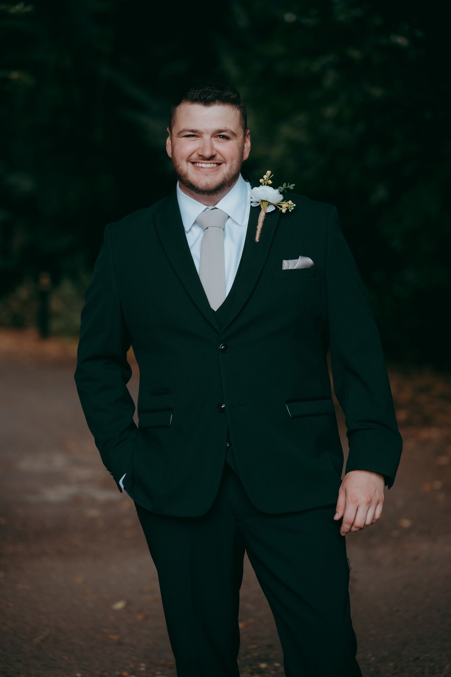 Smiling groom in dark green three-piece suit with white tie and boutonniere posing outdoors at wedding.