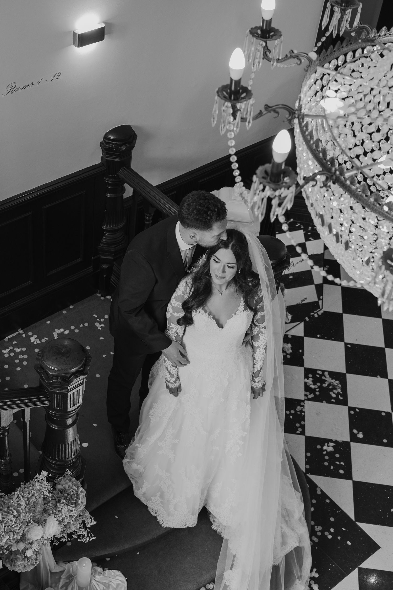 Black and white wedding photo of bride and groom sharing a tender moment on elegant staircase with chandelier.