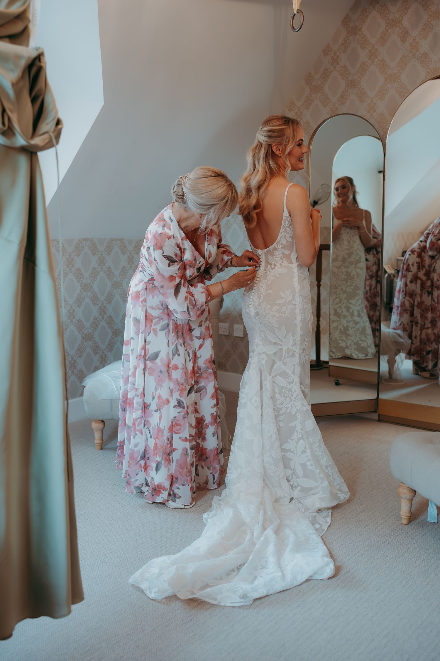 A bride in a lace wedding gown gets help with her dress from a woman in a floral robe in an elegant bridal suite.
