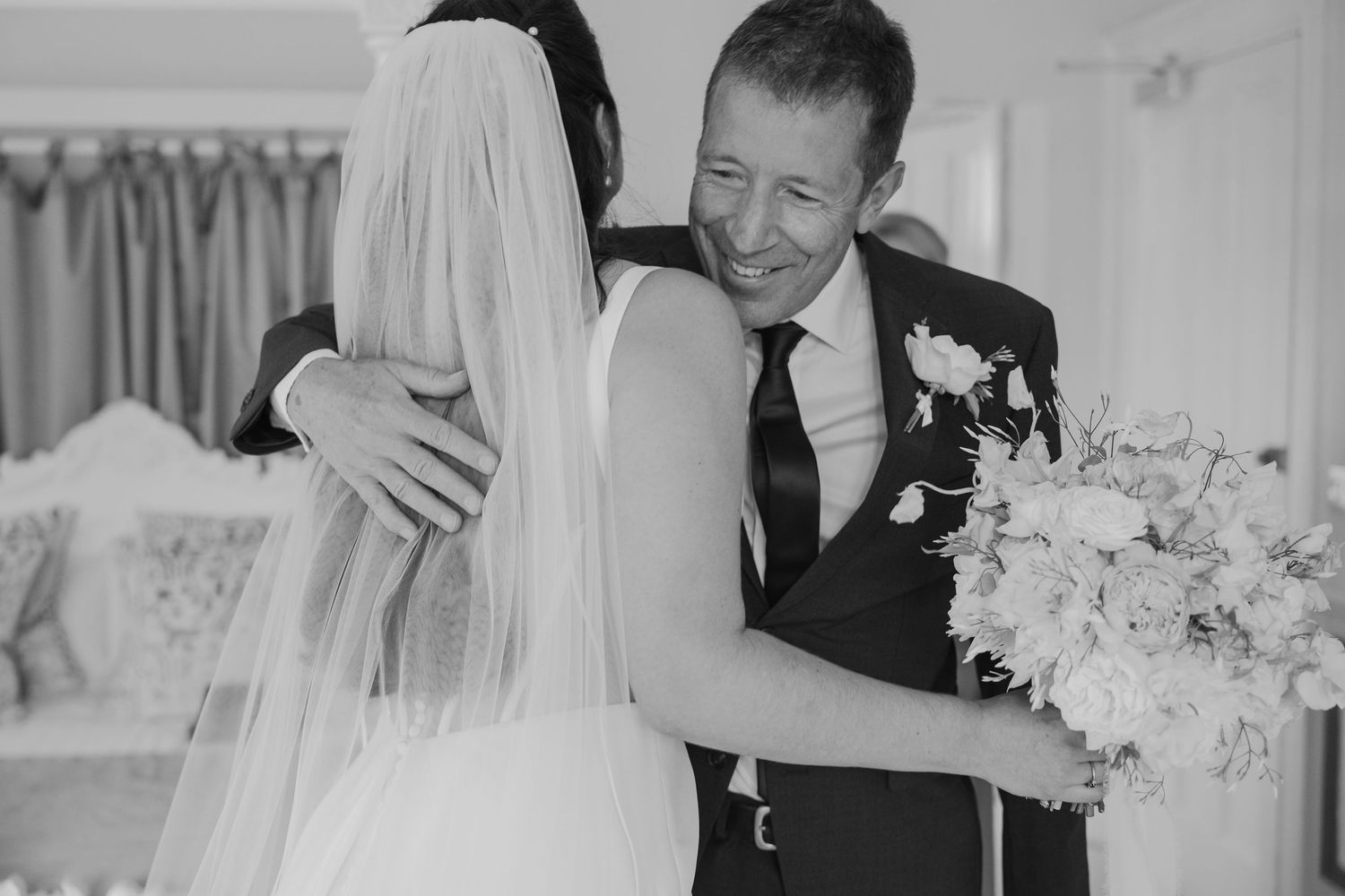 Black and white photo of a groom smiling and embracing a bride holding a bouquet on their wedding day.