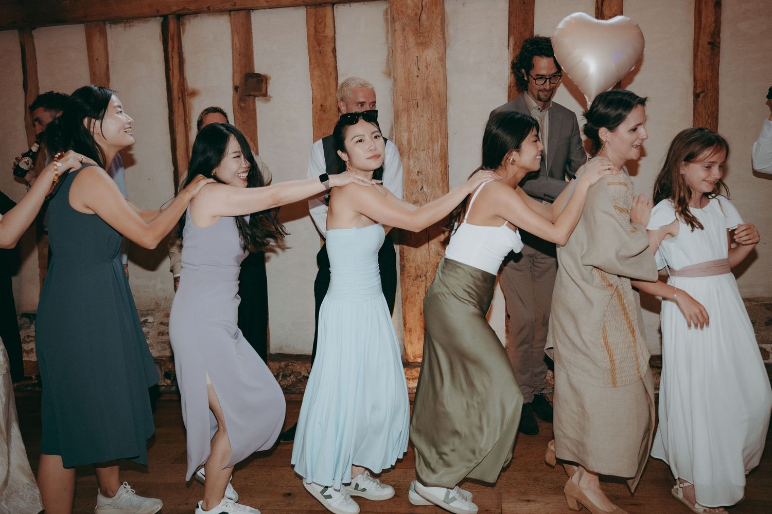 Women in elegant dresses dance in a line at a wedding reception in a rustic venue with wood-paneled walls.