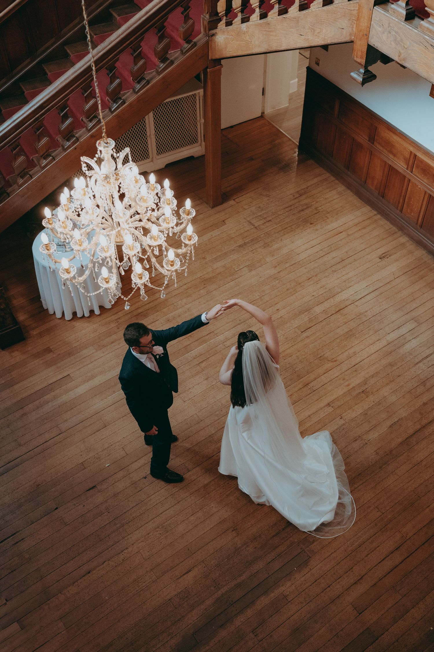 Aerial view of bride and groom sharing first dance under crystal chandelier on wooden ballroom floor.