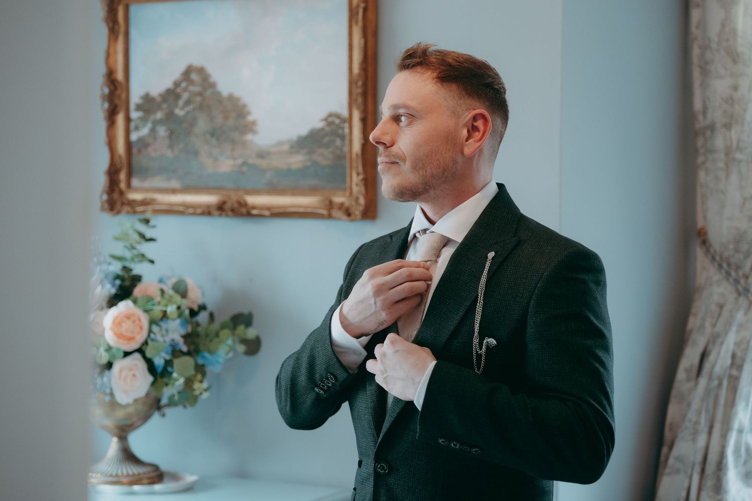 Groom in dark suit adjusting tie before wedding ceremony, standing by floral arrangement and vintage painting.