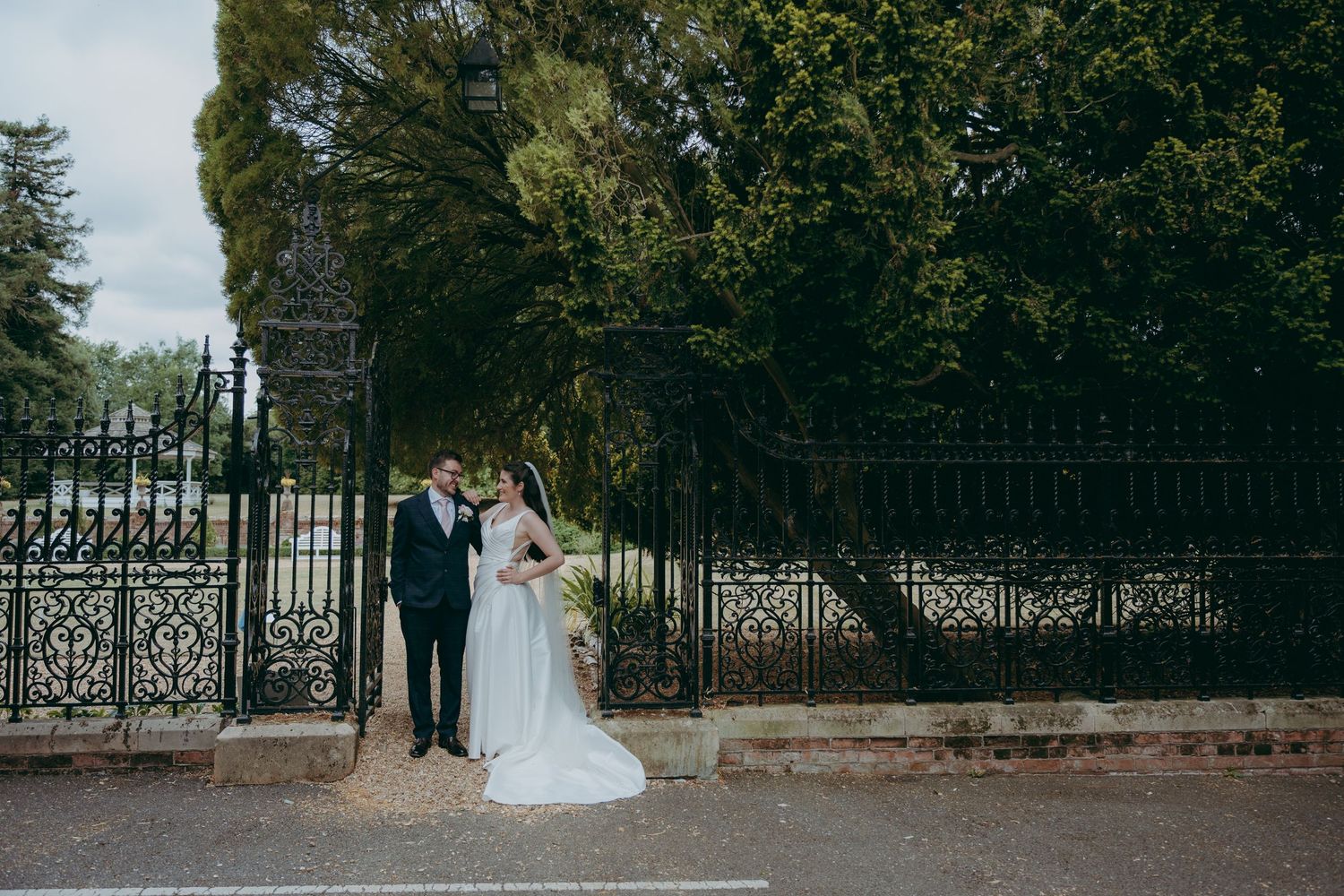 Bride and groom share a romantic moment by ornate iron gates surrounded by lush green trees on their wedding day.