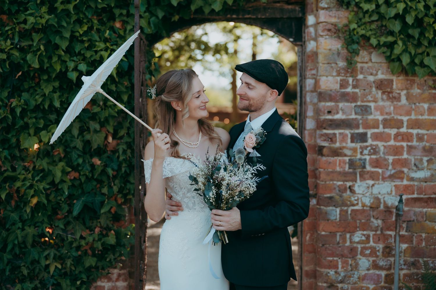 Bride and groom share a romantic moment by a brick archway, she holds a bouquet and parasol in a lace wedding dress.