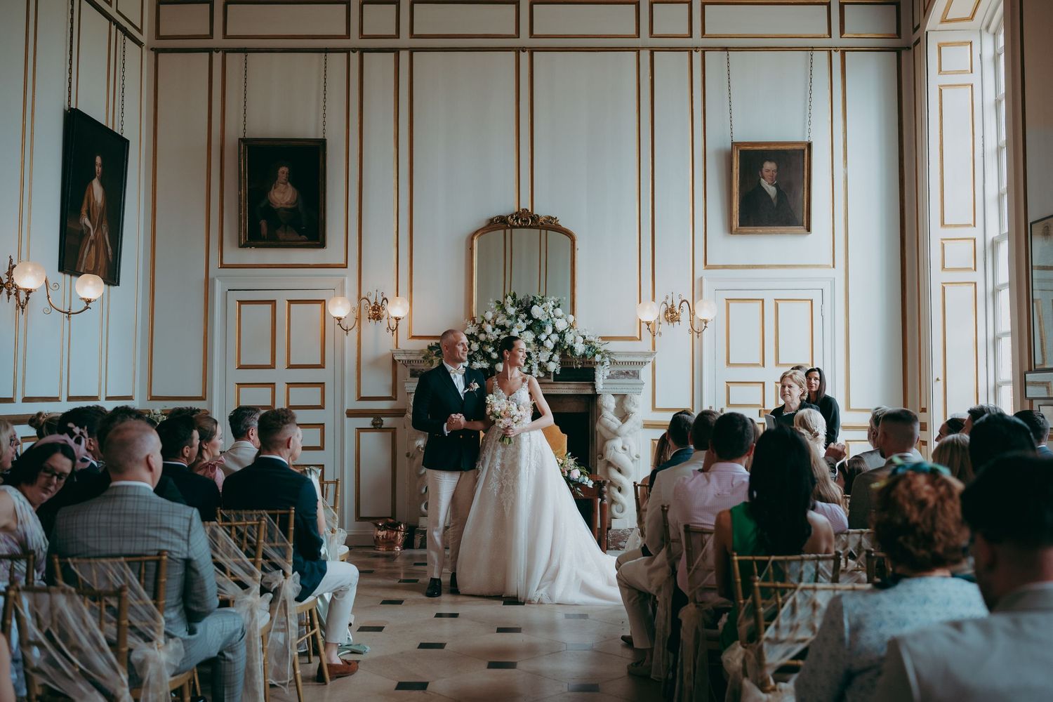Elegant wedding ceremony in a grand white and gold ballroom with bride and groom at the altar surrounded by seated guests.