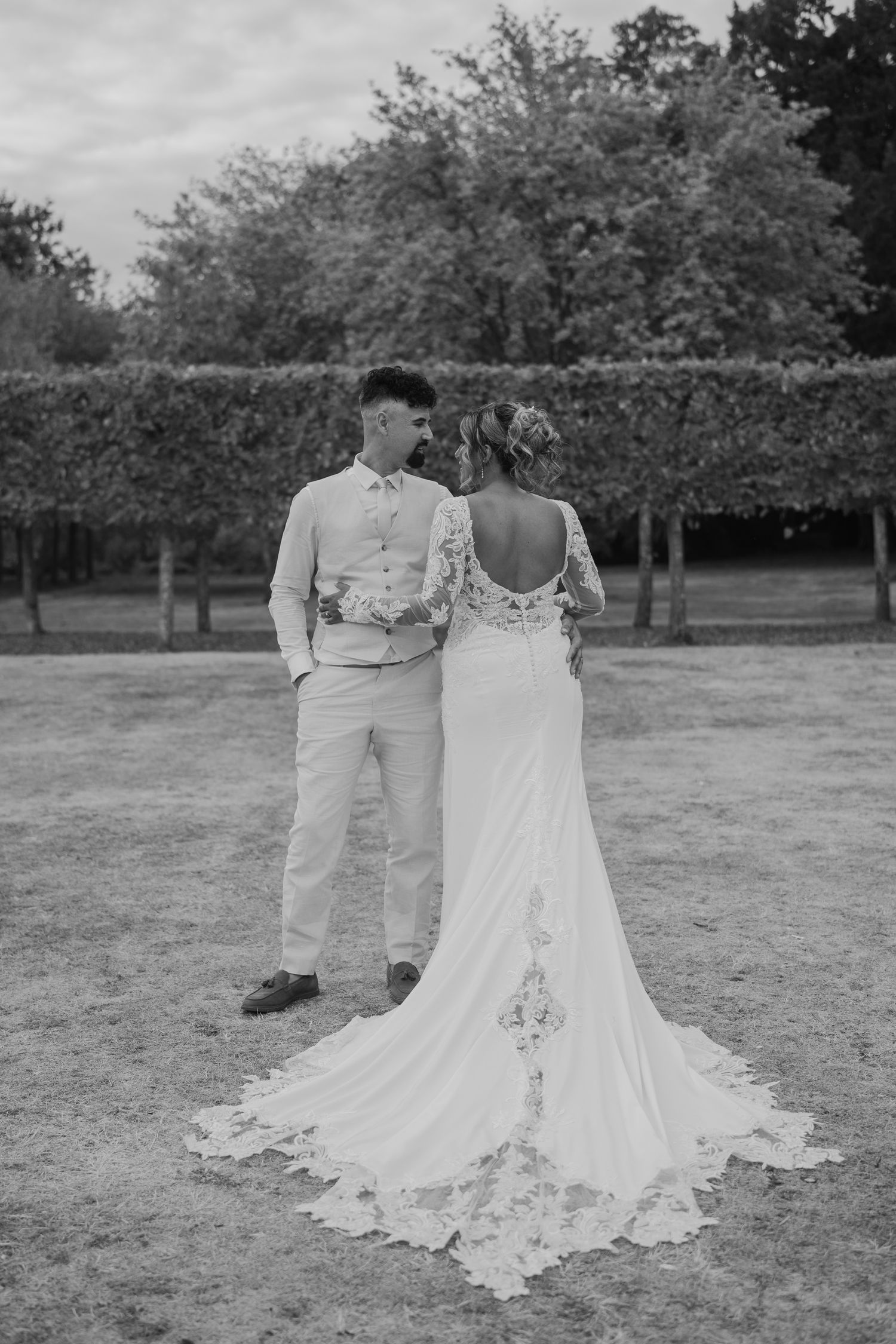 Black and white photo of bride and groom standing together outdoors, bride in lace wedding gown with train.