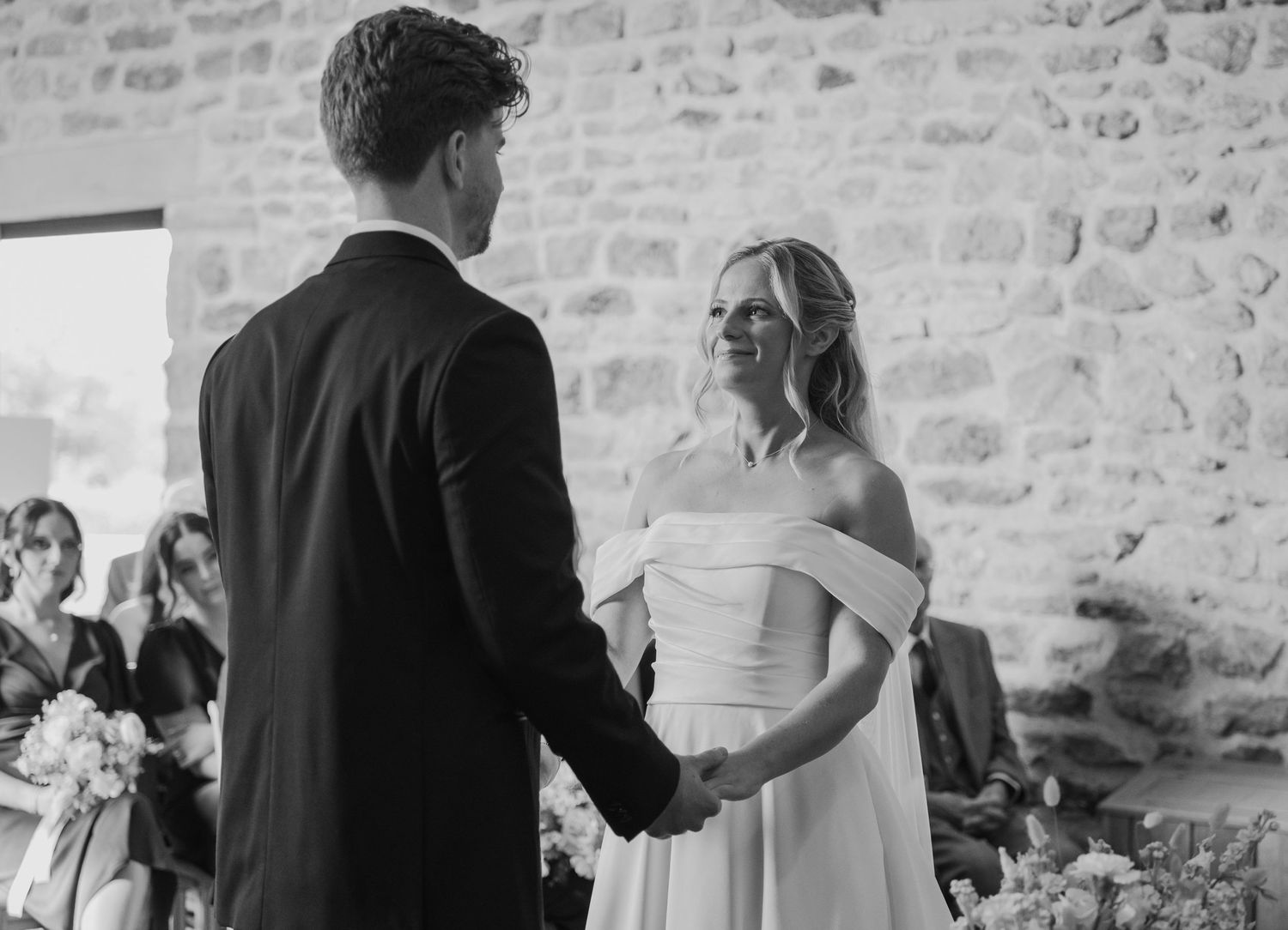 Black and white wedding ceremony photo of bride in off-shoulder gown holding hands with groom in suit at rustic venue.