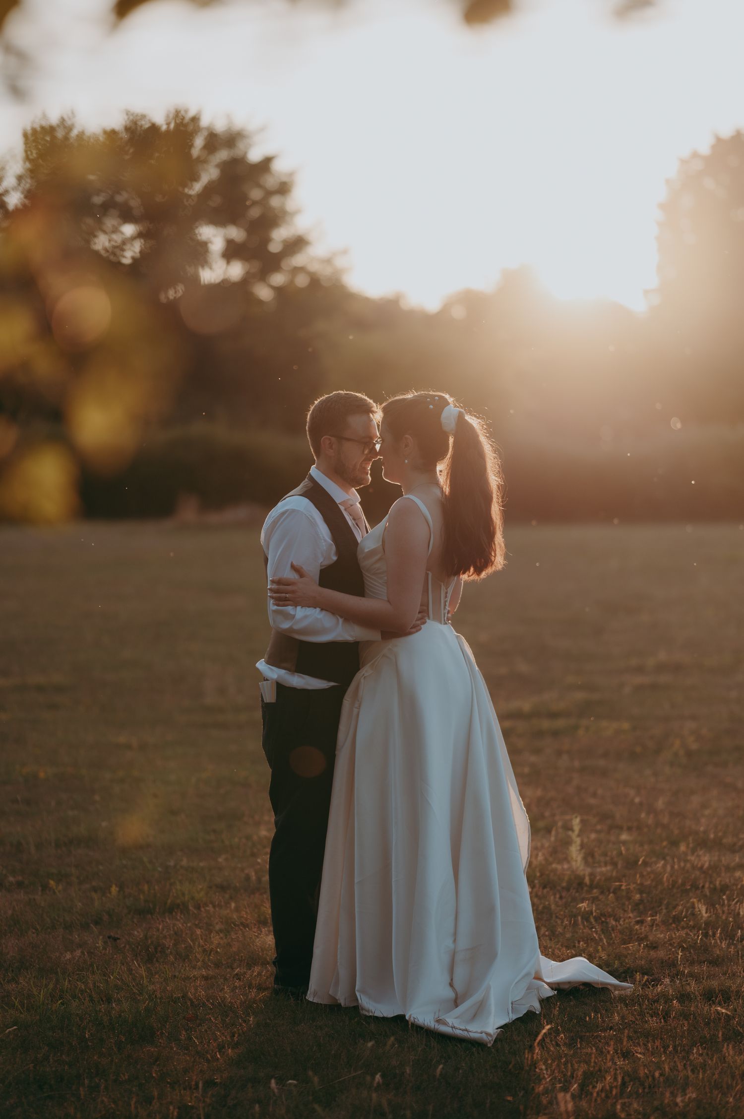 A bride and groom share a romantic kiss at golden hour in an open field, bathed in warm sunset light.