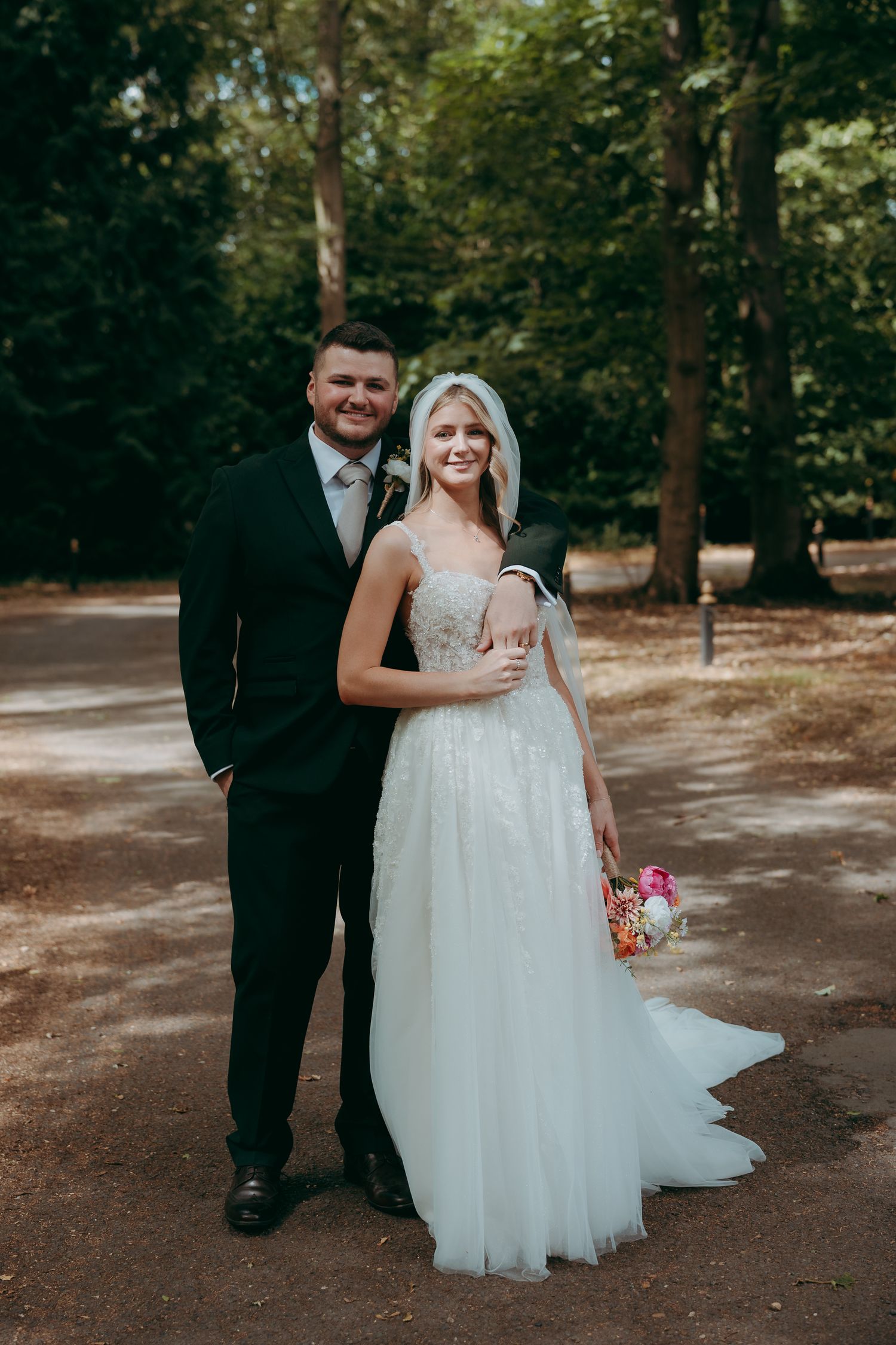 Bride and groom pose together on a wooded path, she in a white gown with bouquet, he in a dark suit.